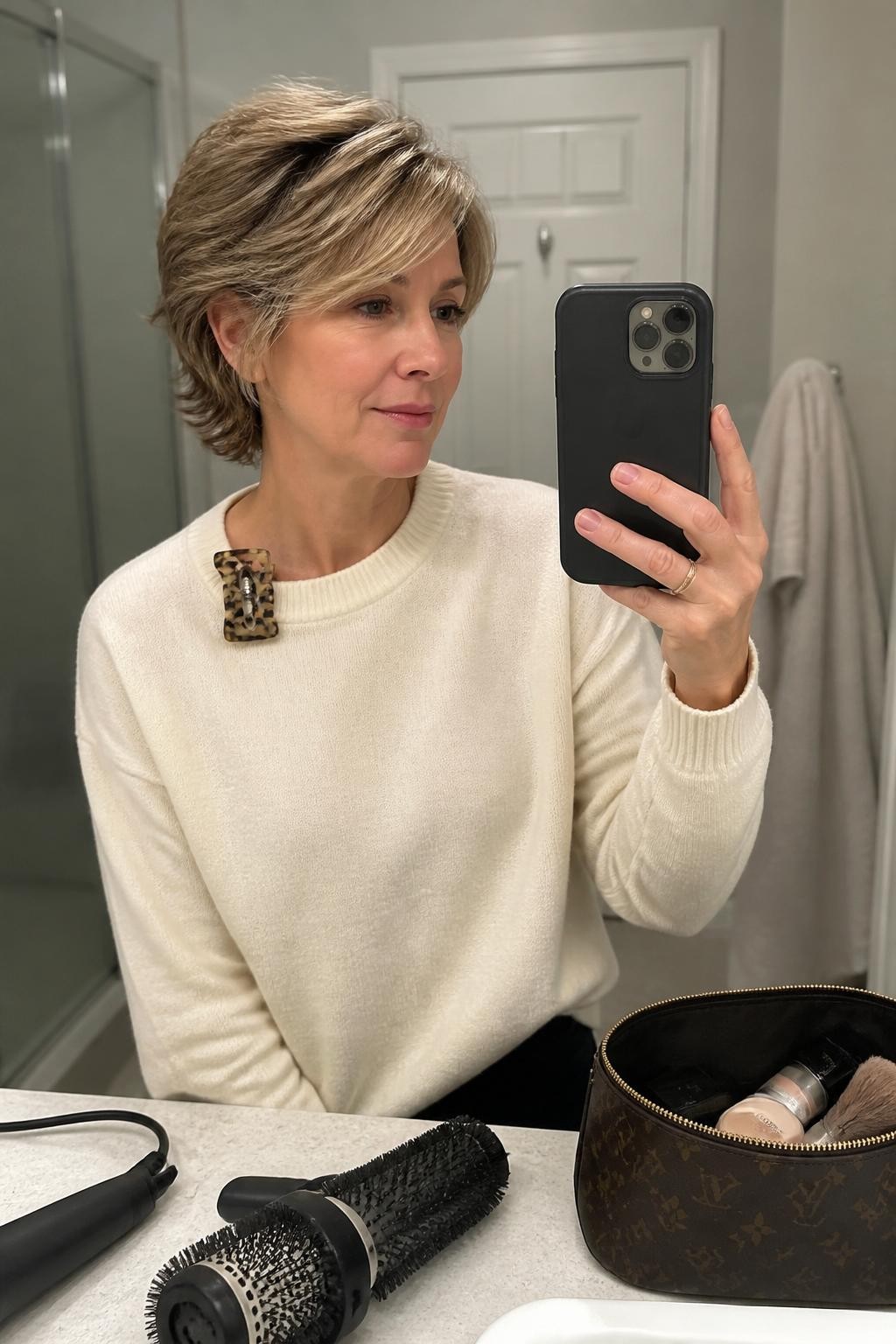 Photo of a woman wearing a layered bixie haircut with airy crown volume, side-swept fringe, and softly flipped ends around the ears and nape, candid mirror selfie in a bathroom vanity mirror, soft indoor lighting, small round brush and blow-dryer on the counter, a tortoiseshell clip attached to her sweater collar and a makeup bag nearby, realistic layered texture, latest iPhone photo quality.