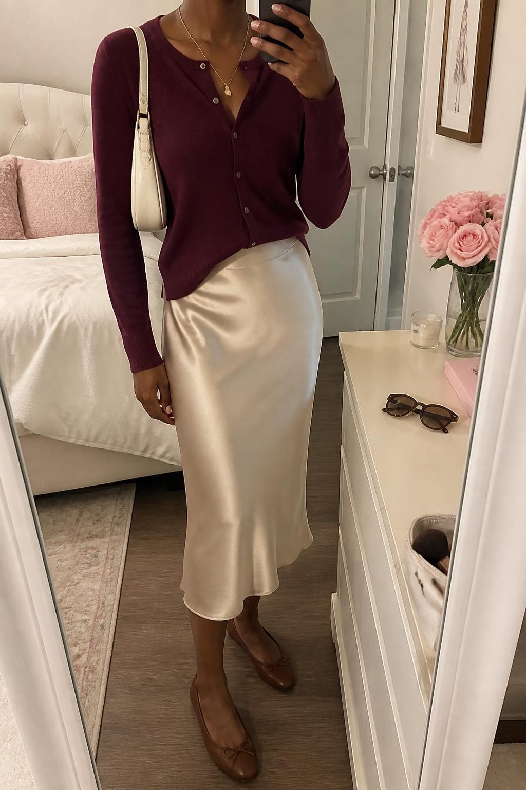 mirror selfie of a Black woman wearing the exact outfit described above, clearly showing a burgundy button-front cardigan tucked into a champagne satin midi slip skirt, brown ballet flats, a cream mini shoulder bag, and delicate gold layered necklaces, holding a phone in front of a mirror, cropped at NECK so the face does not show, with a relaxed casual selfie pose, in a feminine-themed bedroom, soft indoor lighting, with sunglasses placed on a dresser and a makeup brush or cosmetic bag on the counter, latest iPhone photo quality.