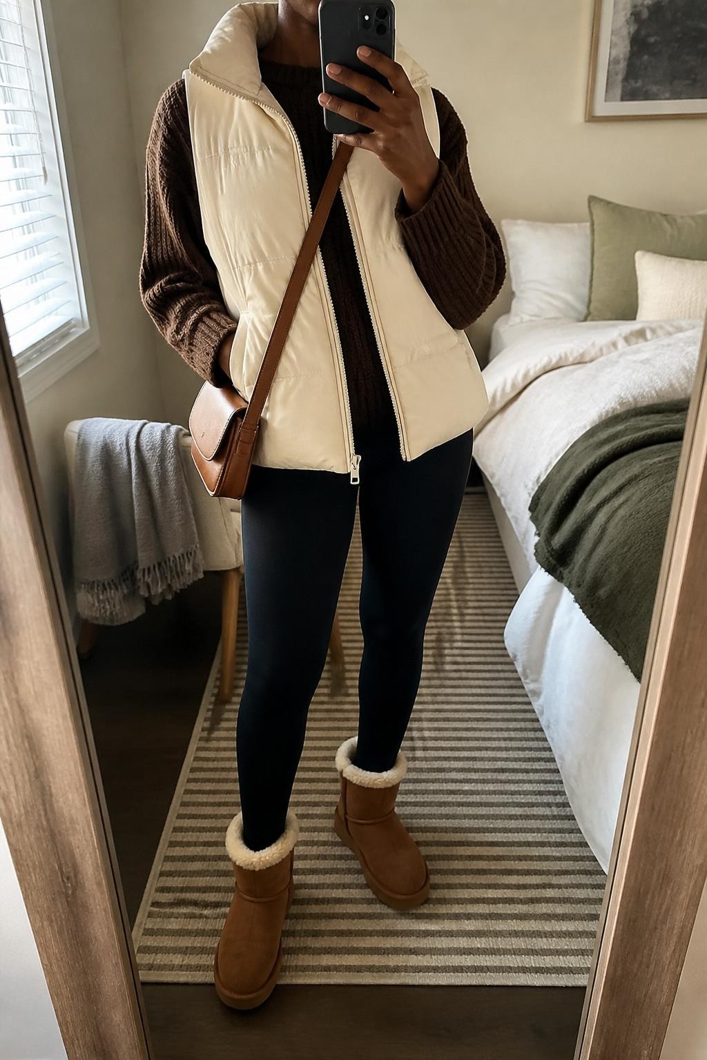 mirror selfie of a Black woman wearing a cream quilted puffer vest over a chocolate brown chunky knit sweater, black leggings, brown shearling-lined boots, and a tan crossbody bag, clearly showing the same clothing items, colors, fabrics, and accessories listed in the Outfit Pieces section, holding a phone in front of a mirror, cropped at NECK so the face does not show, with a relaxed casual selfie pose, in a cozy bedroom mirror setup, natural window light, including soft pillows on an unmade bed and a folded blanket on a chair, latest iPhone photo quality.
