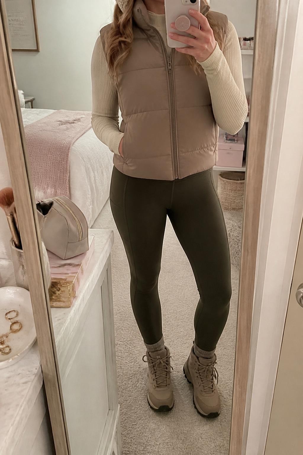 mirror selfie of a woman wearing the exact outfit described above, clearly showing a taupe puffer vest, cream ribbed long-sleeve base layer, dark olive hiking leggings, taupe hiking boots, and a cream knit headband, holding a phone in front of a mirror, cropped at NECK so the face does not show, with a relaxed casual selfie pose, in a feminine-themed bedroom, soft indoor lighting, with a jewelry tray on the vanity and a makeup brush or cosmetic bag on the counter, latest iPhone photo quality.