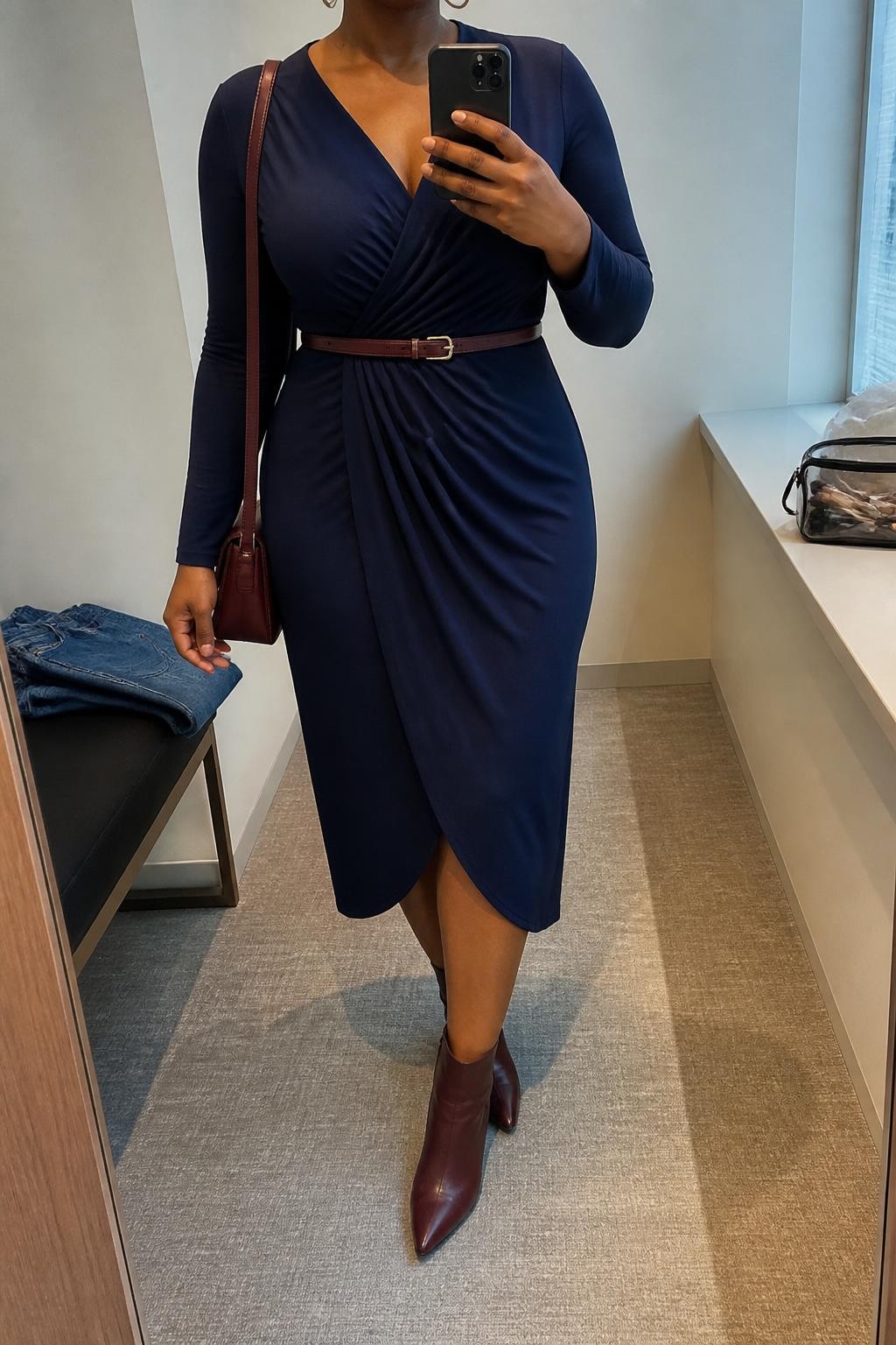 mirror selfie of a Black curvy woman wearing a navy long-sleeve wrap dress, burgundy ankle boots, gold hoop earrings, slim brown belt, and a burgundy shoulder bag, clearly showing the same clothing items, colors, fabrics, and accessories listed in the Outfit Pieces section, holding a phone in front of a mirror, cropped at NECK so the face does not show, with a relaxed casual selfie pose, in a minimal dressing room, natural window light, including folded denim jeans on a bench and a makeup brush or cosmetic bag on the counter, latest iPhone photo quality.