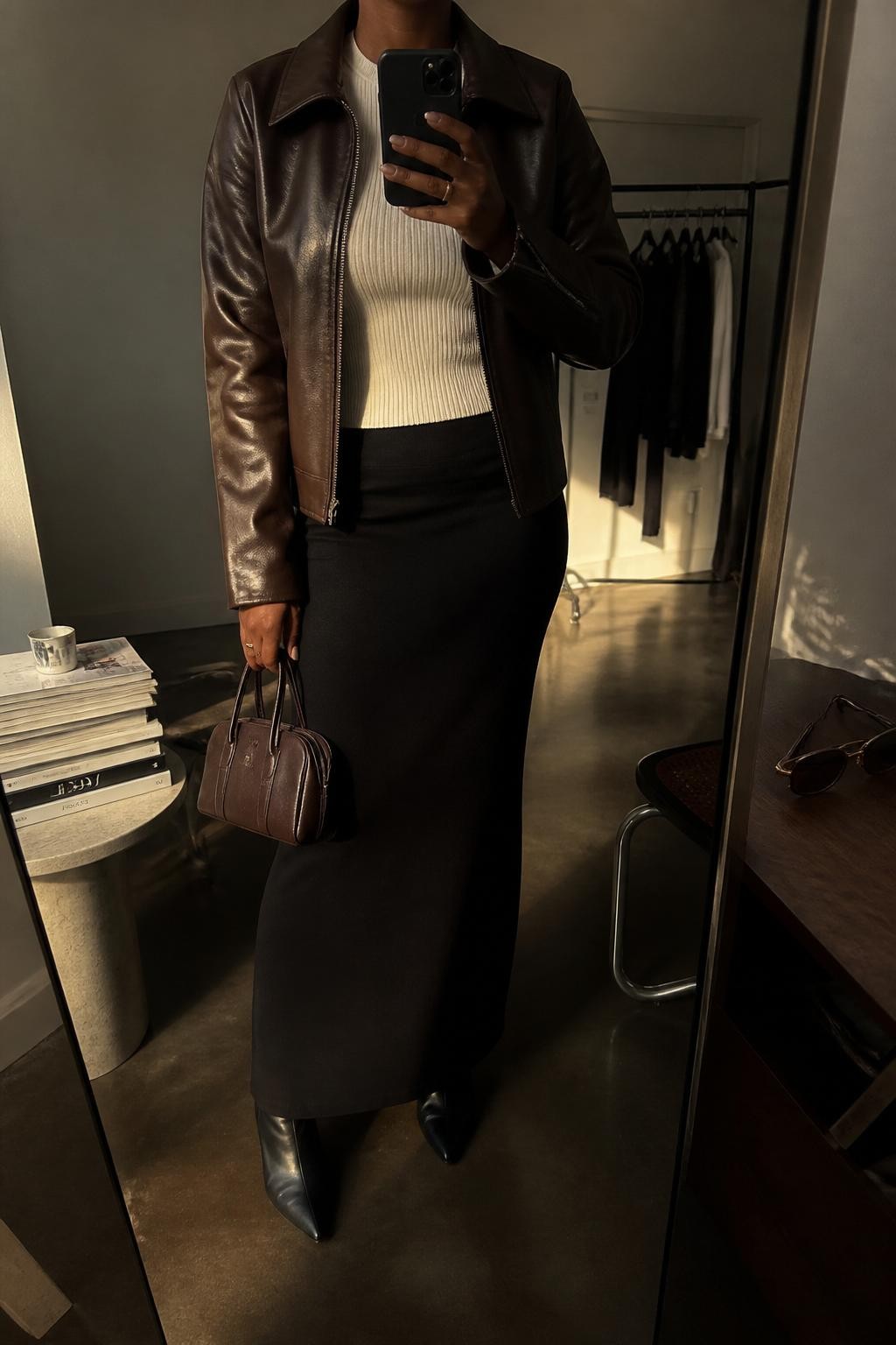 mirror selfie of a Black woman wearing an espresso brown leather jacket over a fitted ivory knit top, black knit maxi skirt, black pointed ankle boots, and an espresso brown mini handbag, clearly showing the same clothing items, colors, fabrics, and accessories listed in the Outfit Pieces section, holding a phone in front of a mirror, cropped at NECK so the face does not show, with a relaxed casual selfie pose, in a fashion studio mirror corner, evening golden hour, including a stack of fashion magazines on a side table and sunglasses placed on a dresser, latest iPhone photo quality.