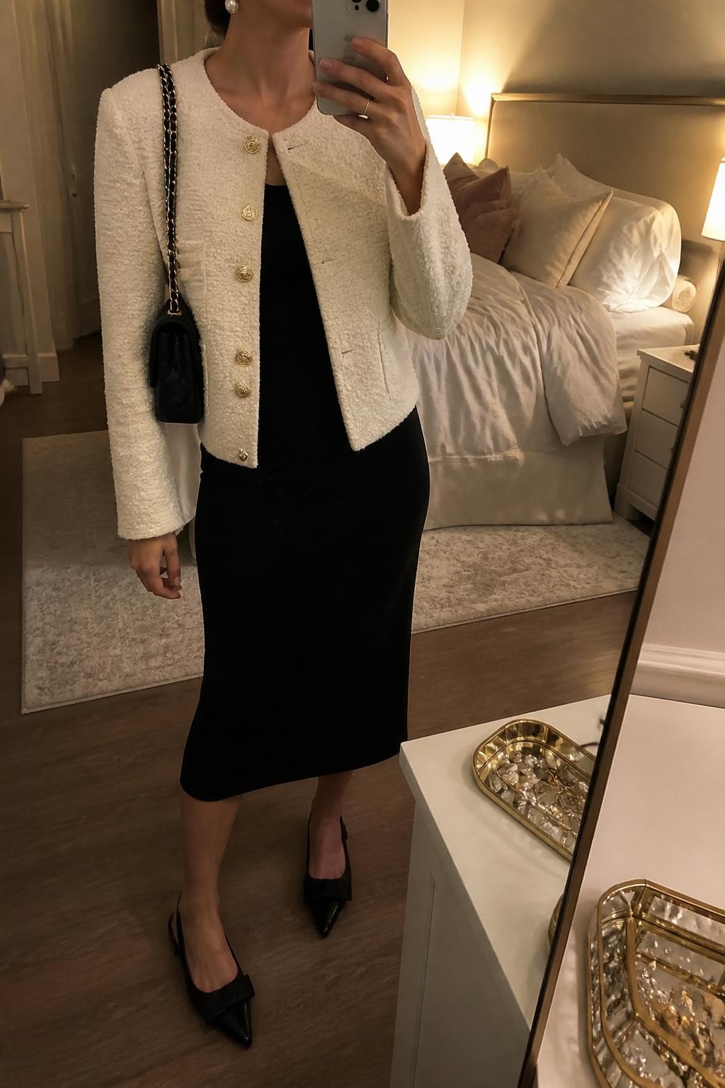 mirror selfie of a woman wearing a cream bouclé jacket, black fitted midi dress, black slingback flats, pearl drop earrings, and a quilted black shoulder bag, clearly showing the same clothing items, colors, fabrics, and accessories listed in the Outfit Pieces section, holding a phone in front of a mirror, cropped at NECK so the face does not show, with a relaxed casual selfie pose, in a feminine-themed bedroom, warm moody lighting, including a jewelry tray on the vanity and soft pillows on an unmade bed, latest iPhone photo quality.