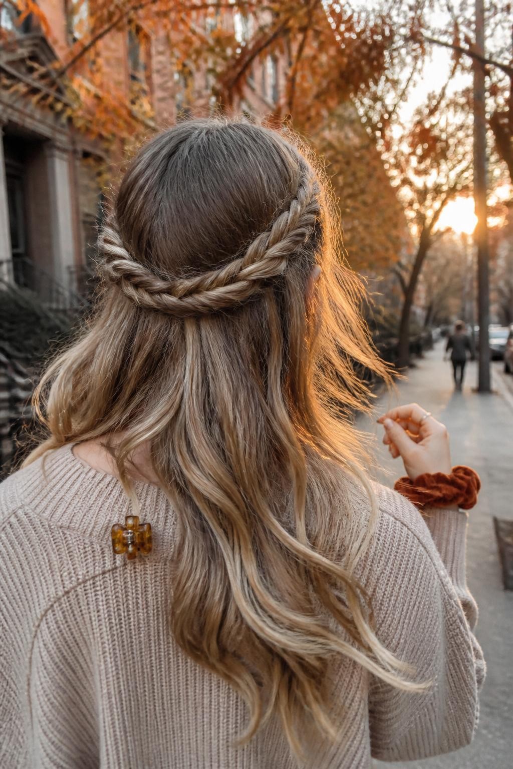 Photo of a woman wearing long loose waves with two medium three-strand braids wrapped around the back of the head to form a soft half-up crown, back-of-head view on a softly blurred city street with copper-colored leaves, golden hour glow, a small amber clip attached to her sweater collar and a velvet scrunchie around her wrist, realistic wisps around the ears and natural wave texture, latest iPhone photo quality.
