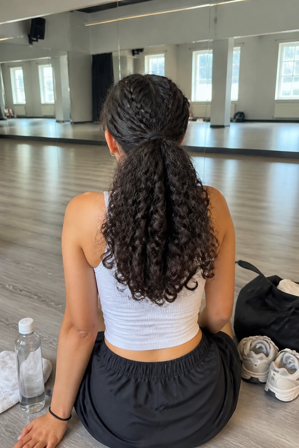 Image prompt
Photo of a woman wearing a half-up curly ponytail with two neat front braids feeding into the top section and full loose curls falling below, back-of-head view inside a bright dance studio, natural window light, containing a small lived-in detail such as a gym towel near a water bottle, a pair of trainers beside a tote bag, and a spare elastic around her wrist, realistic pre-class moment, latest iPhone photo quality.