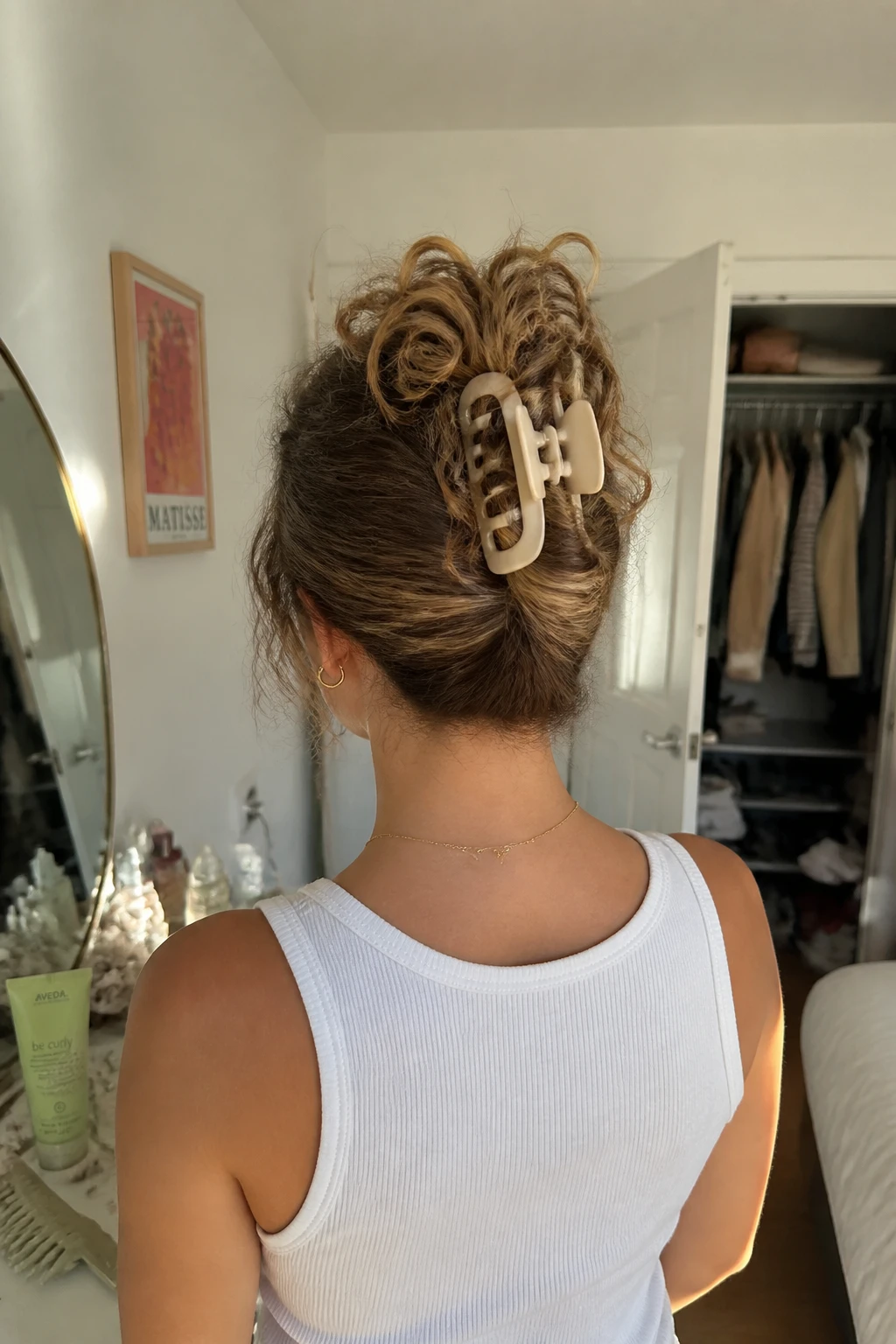 Image prompt

Photo of a woman wearing a curly claw clip updo with a vertical twist and soft curl ends spilling over the top of a large clip, back-of-head view highlighting the clip placement, standing in a sun-drenched bedroom, natural window light, containing a small lived-in detail such as curl cream and a wide-tooth comb on the vanity and a slightly open closet behind her, realistic getting-ready moment, latest iPhone photo quality.