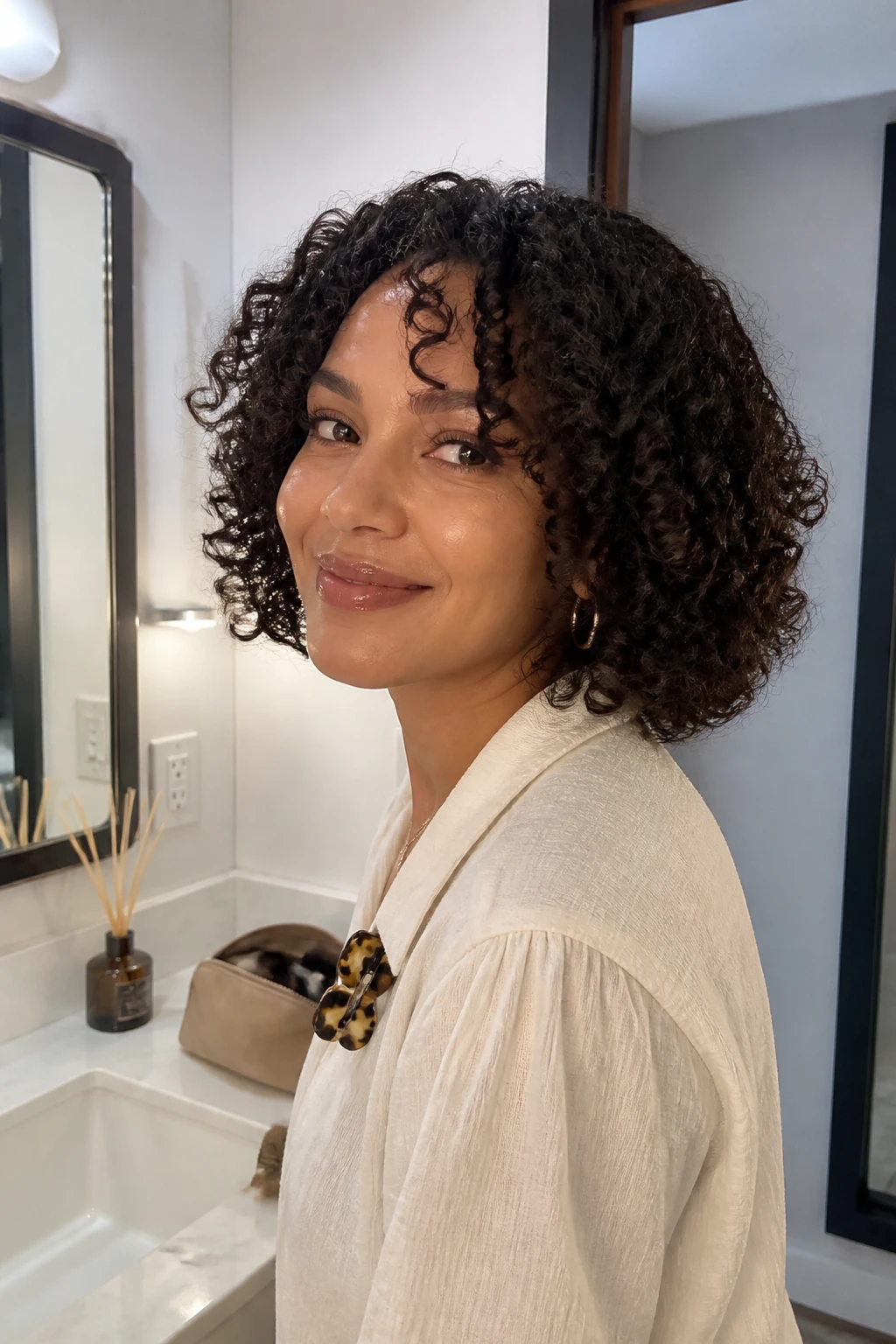 Image prompt
Photo of a woman wearing a chin-length 3B curly bob with soft curtain bangs opening around the forehead, springy ringlets framing the jaw, and gentle volume at the sides, front three-quarter view beside a bathroom vanity mirror, soft indoor lighting, containing a small lived-in detail such as a diffuser on the counter, a makeup bag nearby, and a claw clip clipped to her blouse collar, realistic getting-ready moment, latest iPhone photo quality.