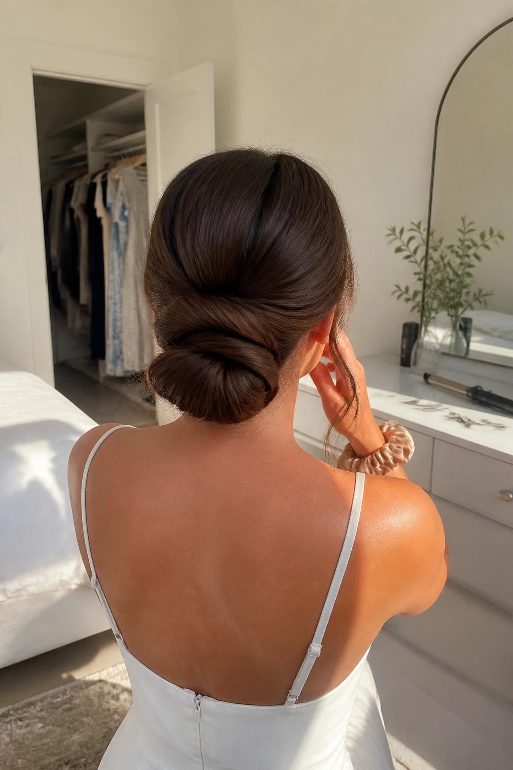 Image prompt: Photo of a woman wearing a low rolled chignon with a softly tucked center, gentle crown volume, and two loose curled side pieces, back-of-head view, sun-drenched bedroom setting, natural window light, bobby pins and a curling iron on the dresser, silk scrunchie around her wrist and an open closet behind her, realistic pre-event getting-ready moment, latest iPhone photo quality.