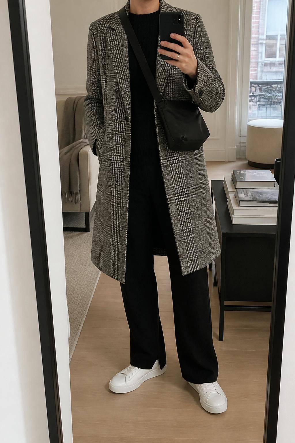 mirror selfie of a woman wearing a gray checked wool coat, black fine-knit crew-neck sweater, black knit straight-leg trousers, white leather sneakers, and a black nylon crossbody bag, clearly showing the same clothing items, colors, fabrics, and accessories listed in the Outfit Pieces section, holding a phone in front of a mirror, cropped at NECK so the face does not show, with a relaxed casual selfie pose, in a stylish city apartment, natural window light, including a stack of fashion magazines on a side table and a folded blanket on a chair, latest iPhone photo quality.
