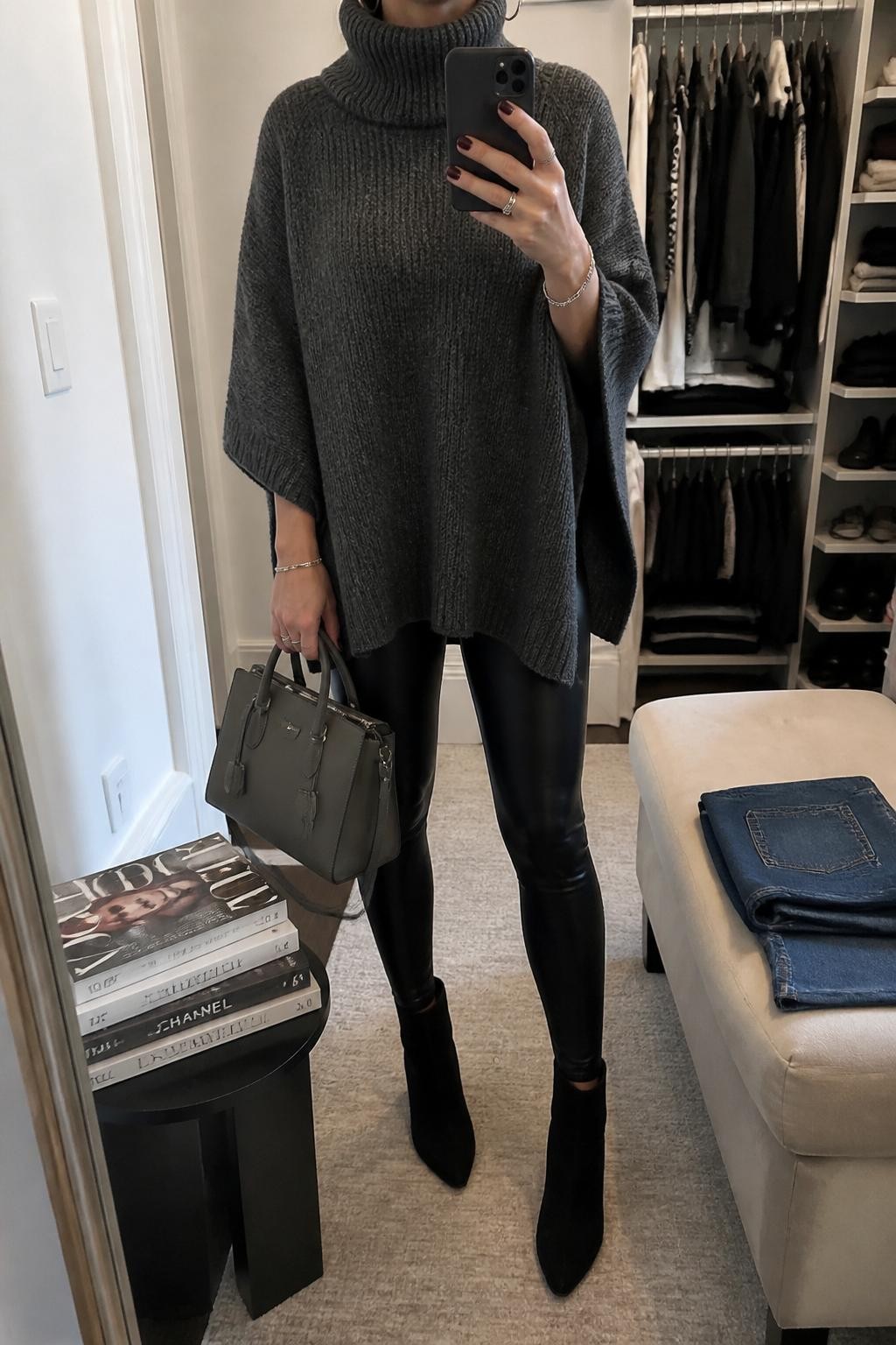 mirror selfie of a woman wearing a charcoal knit turtleneck poncho over black faux leather leggings, black suede ankle boots, silver hoop earrings, and a gray structured handbag, clearly showing the same clothing items, colors, fabrics, and accessories listed in the Outfit Pieces section, holding a phone in front of a mirror, cropped at NECK so the face does not show, with a relaxed casual selfie pose, in a walk-in closet mirror, soft indoor lighting, including a stack of fashion magazines on a side table and folded denim jeans on a bench, latest iPhone photo quality.