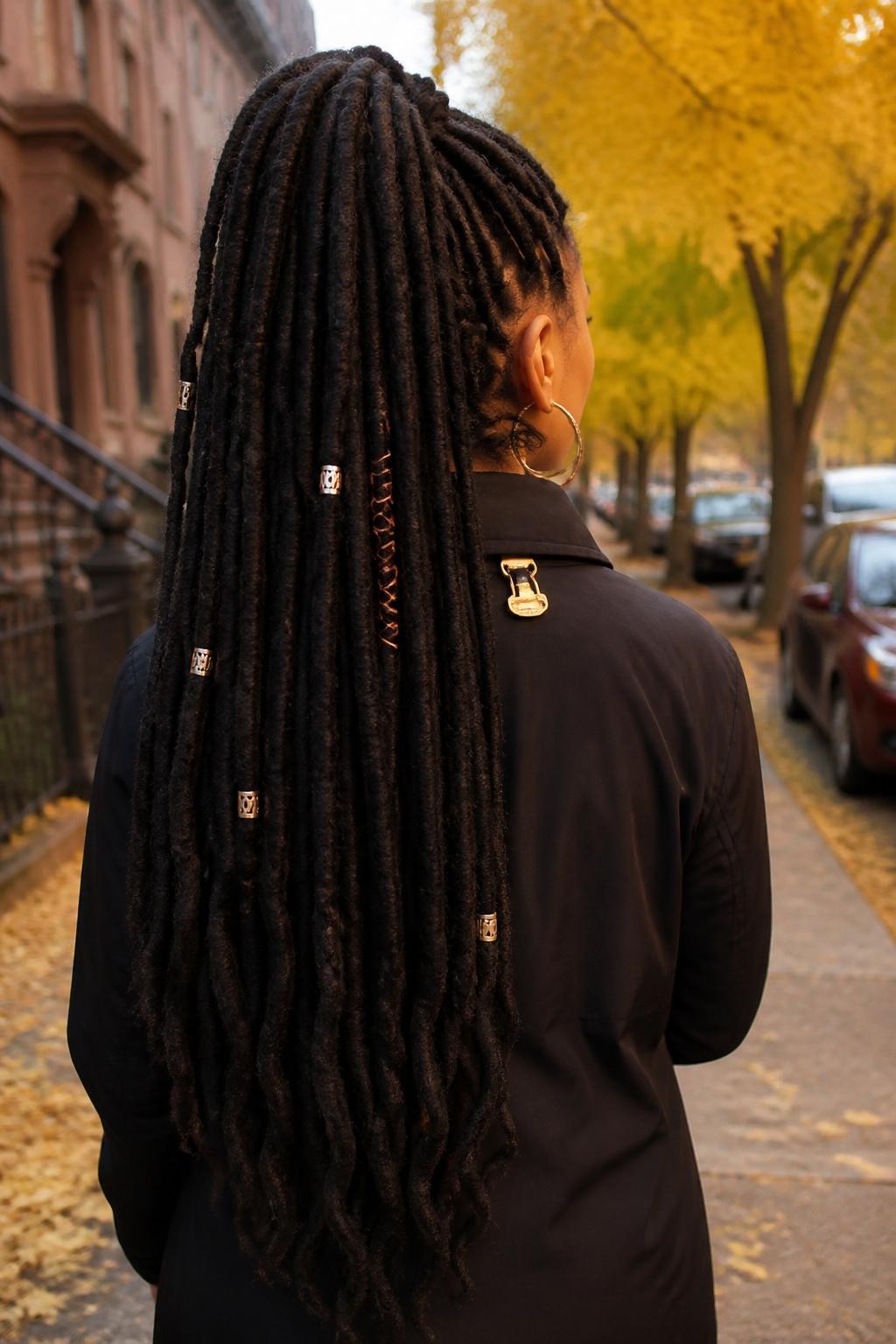 Photo of a Black woman wearing very long soft faux locs styled into a high half-up ponytail with the remaining locs flowing down her back, decorated with scattered copper cuffs and one small rust-colored cord wrap, back-of-head view on a softly blurred city street with golden fall foliage, warm sunset glow, small hair clip attached to her jacket collar and large hoop earrings peeking through, realistic candid autumn portrait, latest iPhone photo quality.