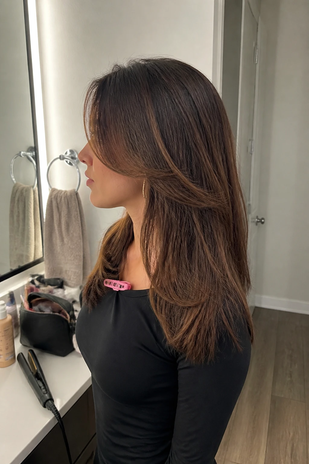 Image prompt
Photo of a woman wearing soft side-part straight hair with smooth face-framing layers and lightly shaped ends, side profile view highlighting the layered pieces, standing at a bathroom vanity mirror with a makeup bag and flat iron on the counter, soft indoor lighting, containing a small lived-in detail such as statement earrings peeking through and a hair clip clipped to her collar, realistic getting-ready moment, latest iPhone photo quality.