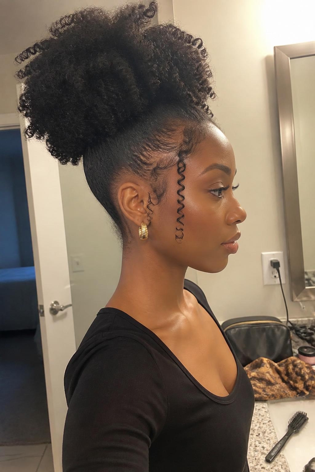 Photo of a Black woman wearing medium-length natural hair styled into a sculpted high puff with full curls, softly laid edges, and two loose spiral tendrils near the temples, side-profile view in a bathroom vanity mirror, warm indoor lighting, edge brush and satin scarf on the counter beside a makeup bag, gold earrings peeking through, realistic natural texture and latest iPhone photo quality.