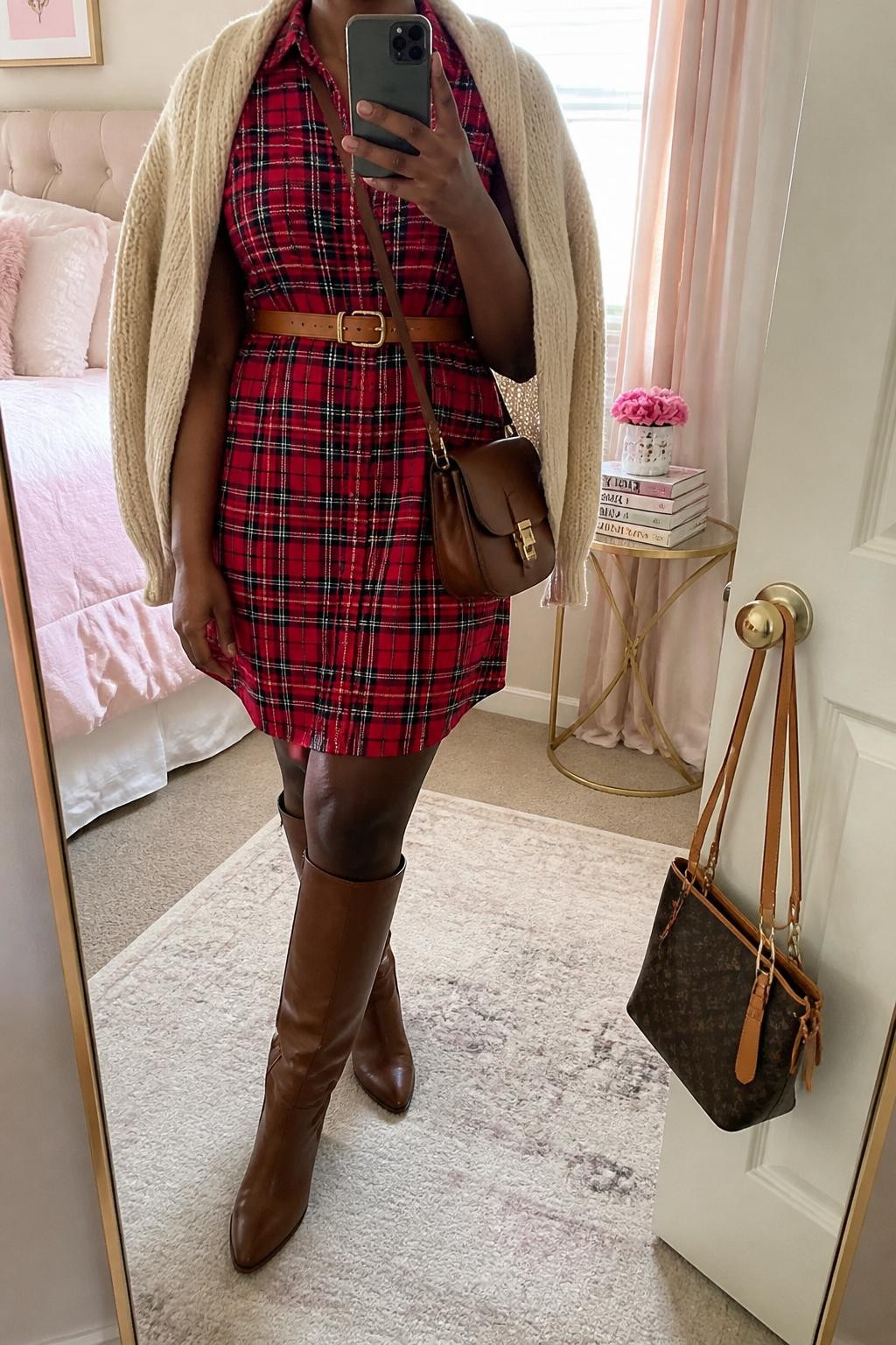 mirror selfie of a Black woman wearing the exact outfit described above, clearly showing a red plaid button-front shirt dress, brown knee-high boots, tan waist belt, cream knit cardigan draped over the shoulders, and a small brown satchel bag, holding a phone in front of a mirror, cropped at NECK so the face does not show, with a relaxed casual selfie pose, in a feminine-themed bedroom, natural window light, with a handbag hanging on a door handle and a stack of fashion magazines on a side table, latest iPhone photo quality.