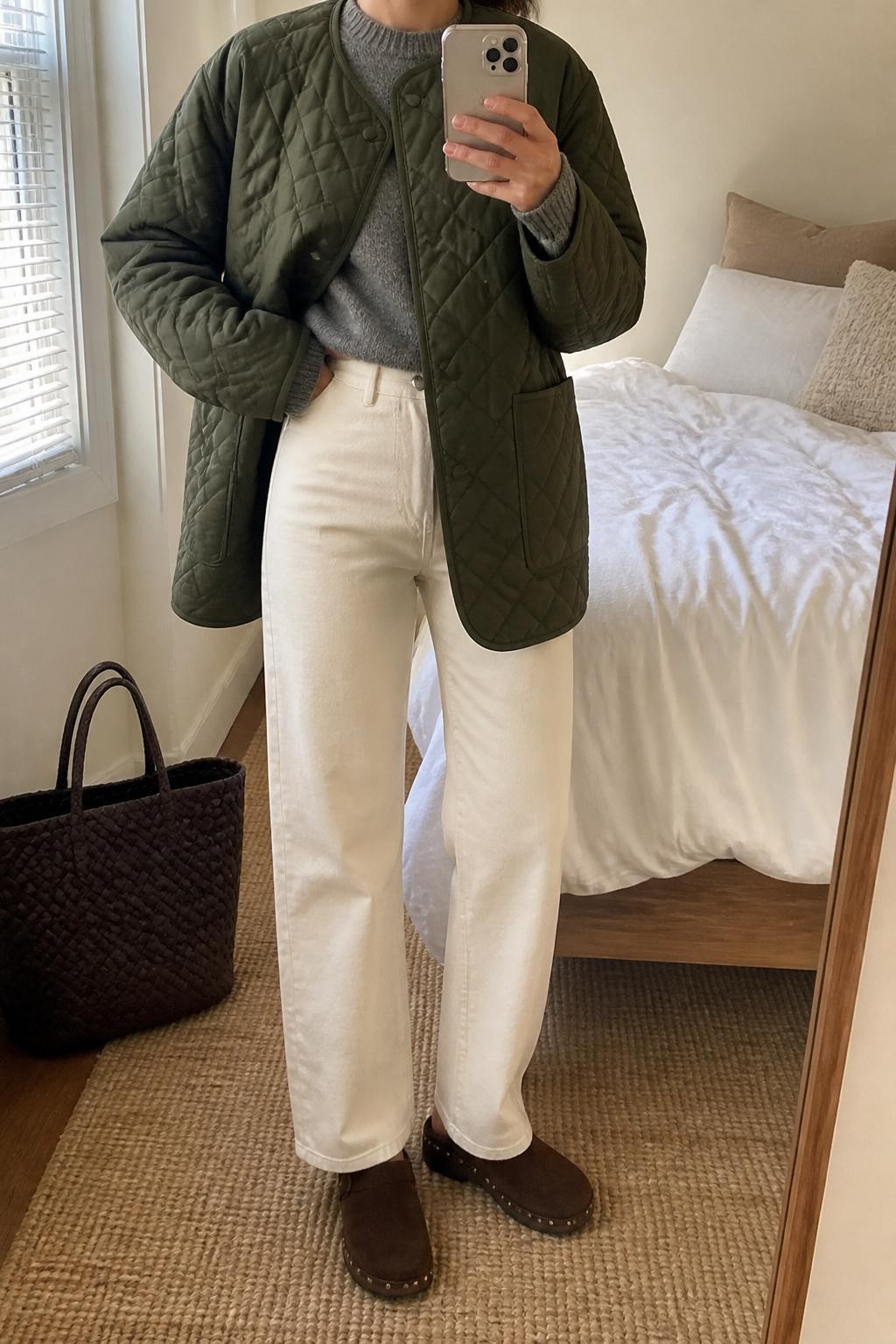 Mirror selfie of a woman wearing an oversized olive green diamond-quilted jacket over a heather gray cashmere crew-neck sweater, high-waisted cream straight-leg jeans, dark brown suede clogs, and a woven dark brown leather tote bag, clearly showing the exact clothing items, colors, fabrics, and accessories listed, holding a latest-model iPhone in front of a mirror, cropped at the neck so the face does not show, relaxed casual selfie pose, in a cozy bedroom mirror setup with natural window light, a tote bag resting on the floor and soft pillows on an unmade bed, natural casual composition, latest iPhone photo quality.
