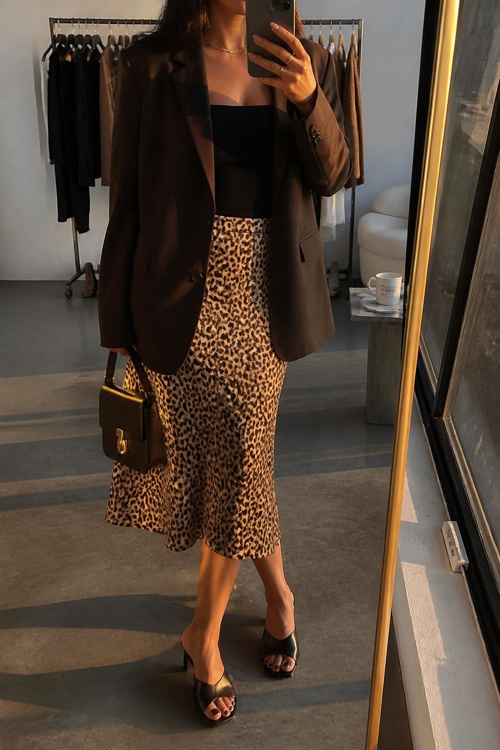 mirror selfie of a Latina woman wearing an espresso brown blazer, black fitted camisole, leopard print midi skirt, black heeled mules, and a small espresso brown handbag, holding a phone in front of a mirror, cropped at NECK so the face does not show, relaxed casual selfie pose, in a fashion studio mirror corner with evening golden hour lighting, including a clothing rack with garments behind and a coffee mug on a side table, realistic latest iPhone photo quality.