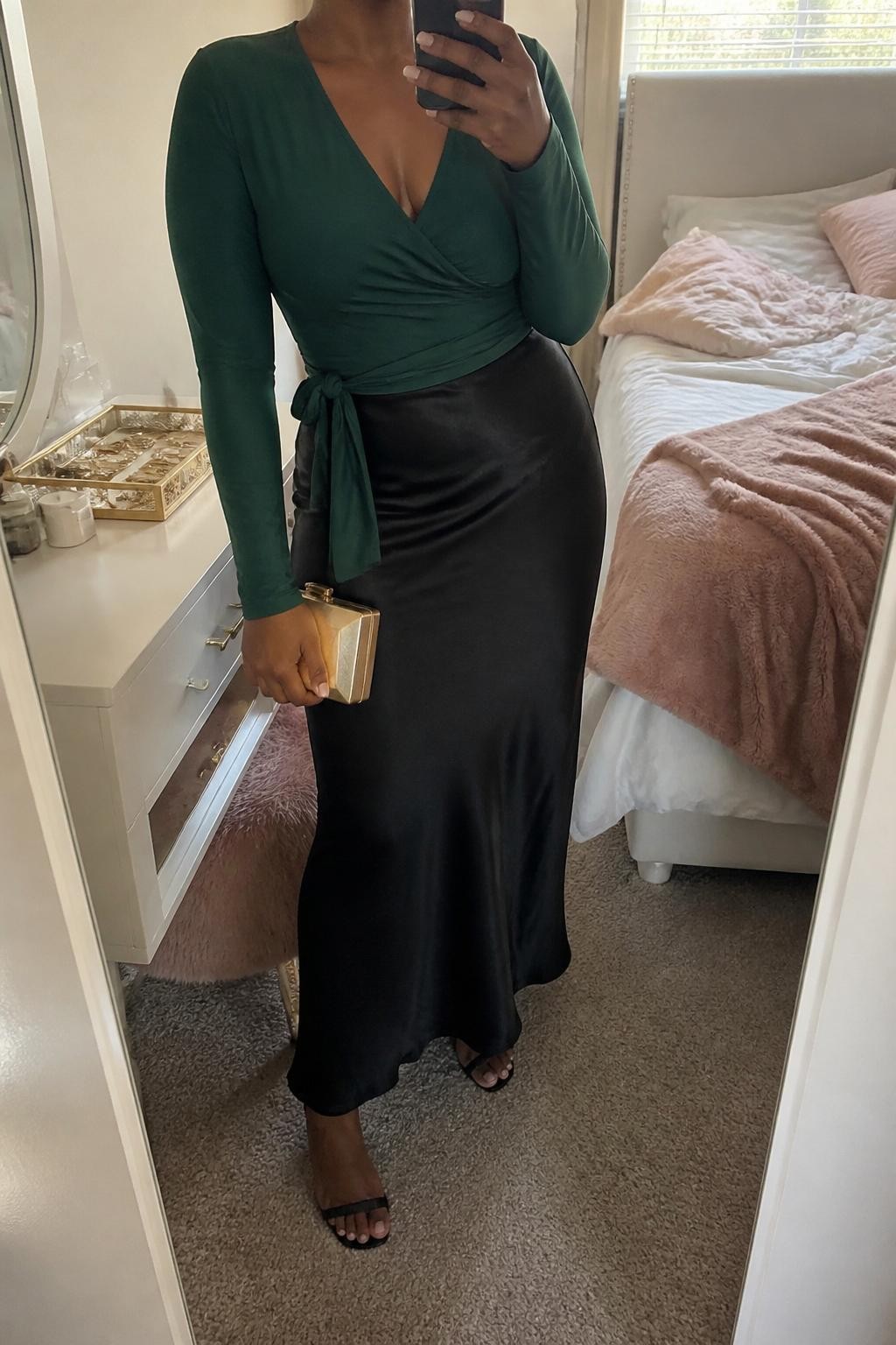 mirror selfie of a Black woman wearing the exact outfit described above, clearly showing a forest green wrap top with long sleeves, black satin maxi skirt, black strappy heels, a gold box clutch, and emerald stud earrings, holding a phone in front of a mirror, cropped at NECK so the face does not show, with a relaxed casual selfie pose, in a feminine-themed bedroom, natural window light, with a jewelry tray on the vanity and soft pillows on an unmade bed, latest iPhone photo quality.