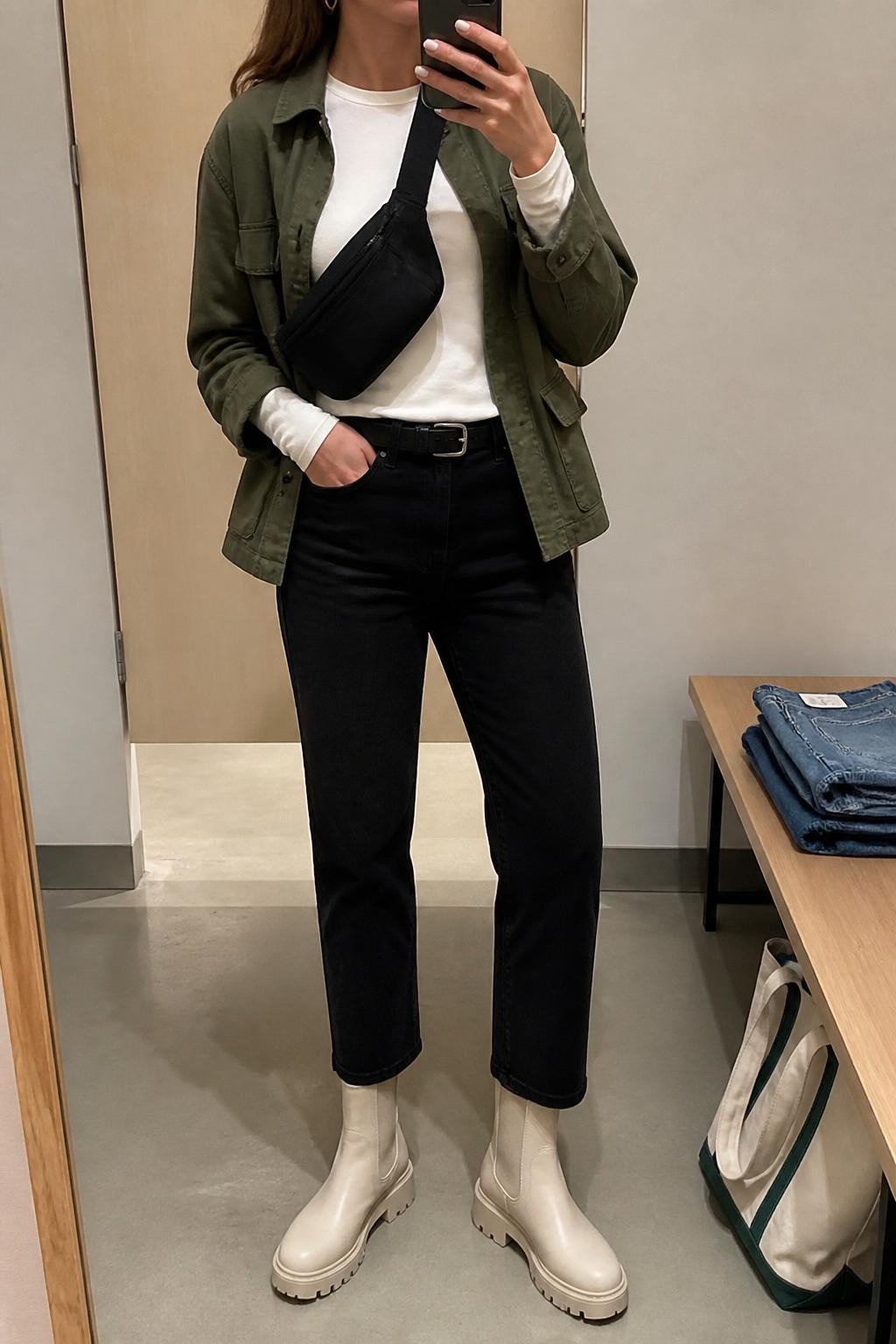 mirror selfie of a woman wearing the exact outfit described above, clearly showing cream Chelsea boots with lug soles, black straight-leg jeans, a fitted white long-sleeve tee, an olive utility jacket, a black belt bag worn crossbody, and small silver hoops, holding a phone in front of a mirror, cropped at NECK so the face does not show, with a relaxed casual selfie pose, in a minimal dressing room, soft indoor lighting, with folded denim jeans on a bench and a tote bag resting on the floor, latest iPhone photo quality.