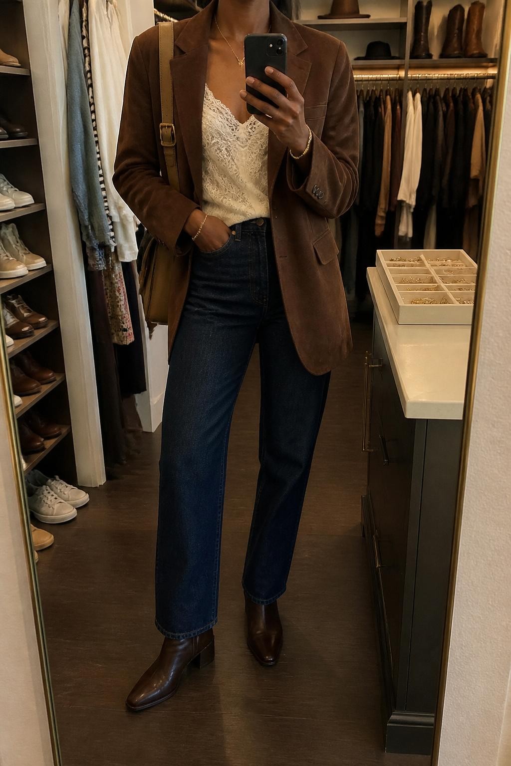 mirror selfie of a Black woman wearing the exact outfit described above, clearly showing a chocolate brown suede blazer, cream lace cami, dark blue straight-leg jeans, chocolate ankle boots, and a small tan shoulder bag, holding a phone in front of a mirror, cropped at NECK so the face does not show, with a relaxed casual selfie pose, in a walk-in closet mirror, warm moody lighting, with a jewelry tray on the vanity and an open closet with clothes visible, latest iPhone photo quality.