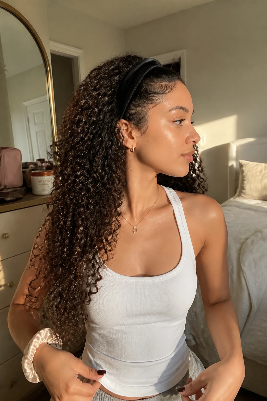 Photo of a woman wearing long defined curls with a deep side part and a slim padded velvet headband positioned behind the hairline, side profile view in a sun-drenched bedroom, natural window light, containing a small lived-in detail such as a curl cream jar on the dresser, a makeup bag nearby, and a silk scrunchie on her wrist, realistic getting-ready moment, latest iPhone photo quality.