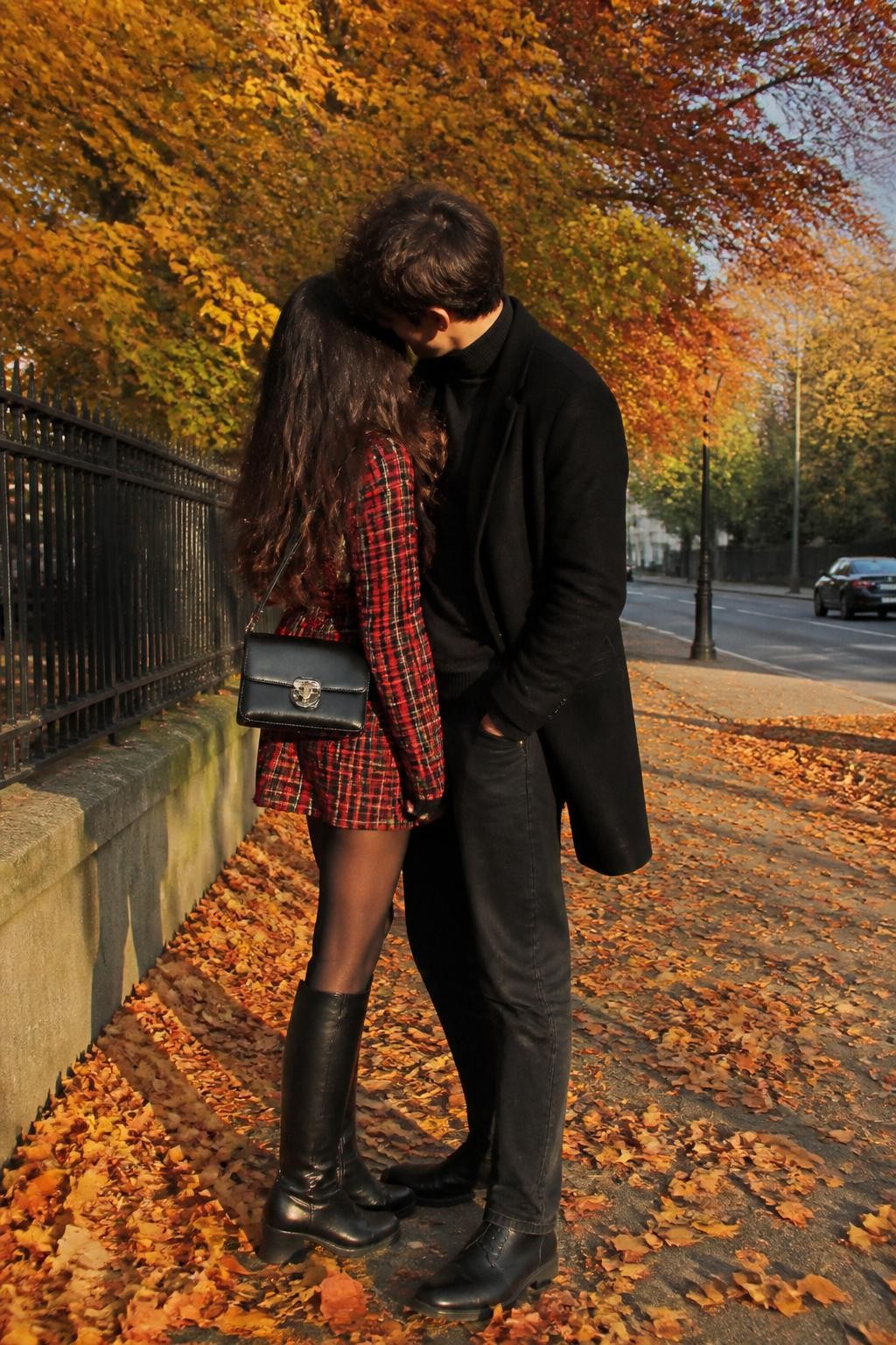 outdoor DSLR photo of a couple wearing romantic fall photoshoot outfits, the woman wearing a red plaid long-sleeve mini dress, sheer black tights, black knee-high boots, and a black mini shoulder bag, the man wearing a black wool coat, charcoal turtleneck, dark jeans, and black Chelsea boots, standing close together on a leaf-covered sidewalk, golden fall trees behind them, warm afternoon lighting, playful candid pose, faces partly hidden by hair and camera angle, realistic autumn couple photography, latest iPhone photo quality.