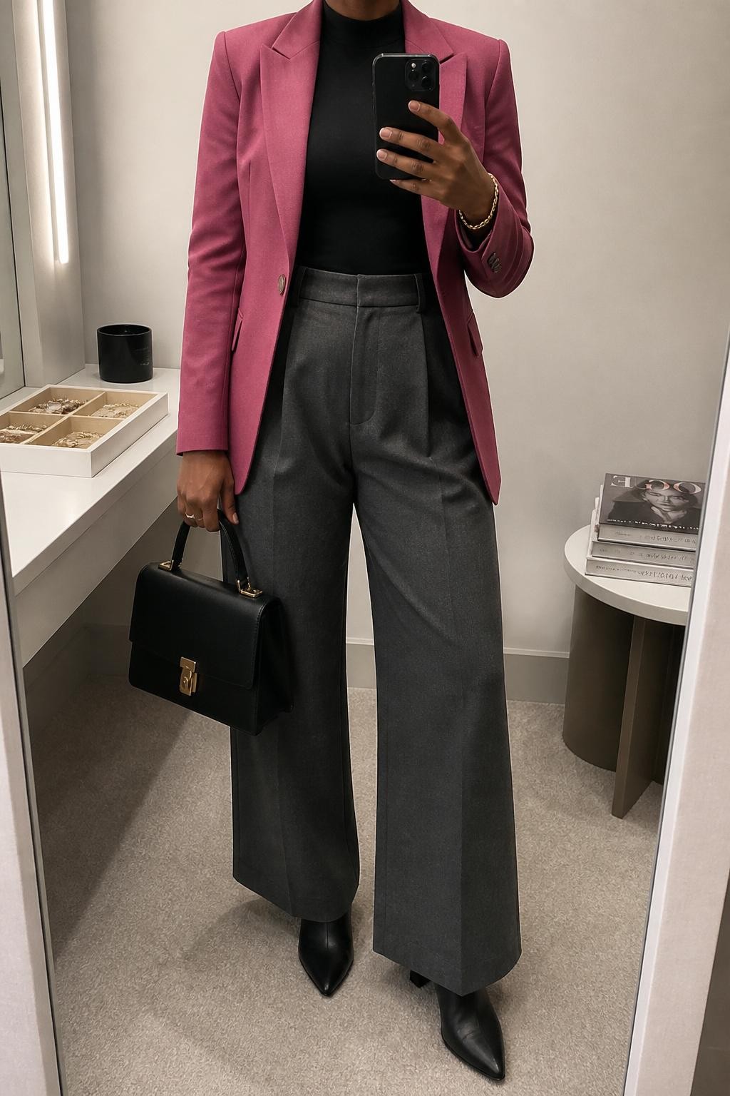 mirror selfie of a Black woman wearing a berry pink tailored blazer over a fitted black mock-neck top, charcoal gray high-waisted wide-leg trousers, black pointed leather ankle boots, and a structured black top-handle bag, clearly showing the same clothing items, colors, fabrics, and accessories listed in the Outfit Pieces section, holding a phone in front of a mirror, cropped at NECK so the face does not show, with a relaxed casual selfie pose, in a minimal dressing room, soft indoor lighting, including a jewelry tray on the vanity and a stack of fashion magazines on a side table, latest iPhone photo quality.