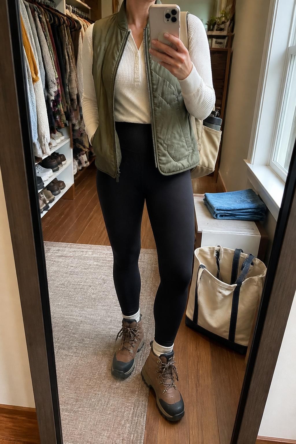 mirror selfie of a woman wearing the exact outfit described above, clearly showing an olive green quilted vest layered over a cream thermal long-sleeve top, black high-waisted trail leggings, brown waterproof hiking boots, and a tan mini hiking backpack, holding a phone in front of a mirror, cropped at NECK so the face does not show, with a relaxed casual selfie pose, in a walk-in closet mirror, natural window light, with a tote bag resting on the floor and folded denim jeans on a bench, latest iPhone photo quality.