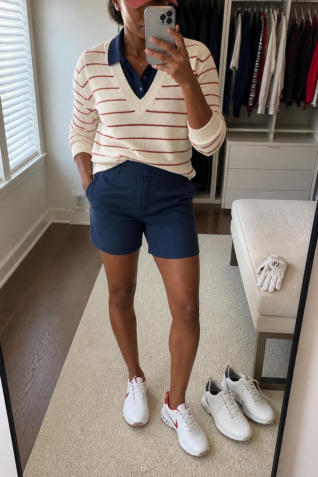 Mirror selfie of a Black woman with warm brown skin wearing a cream knit sweater with thin brick-red horizontal stripes over a navy sleeveless collared golf polo, navy tailored mid-thigh golf shorts, white spikeless golf shoes with red detailing, and a brick-red baseball cap, clearly showing the exact clothing items, colors, stripes, fabrics, and accessories listed, holding a latest-model iPhone in front of a mirror, cropped at the neck so the face does not show, relaxed casual selfie pose, in a walk-in closet mirror with natural window light, a pair of golf shoes on the floor and a golf glove resting on a bench, natural uncluttered composition, latest iPhone photo quality.