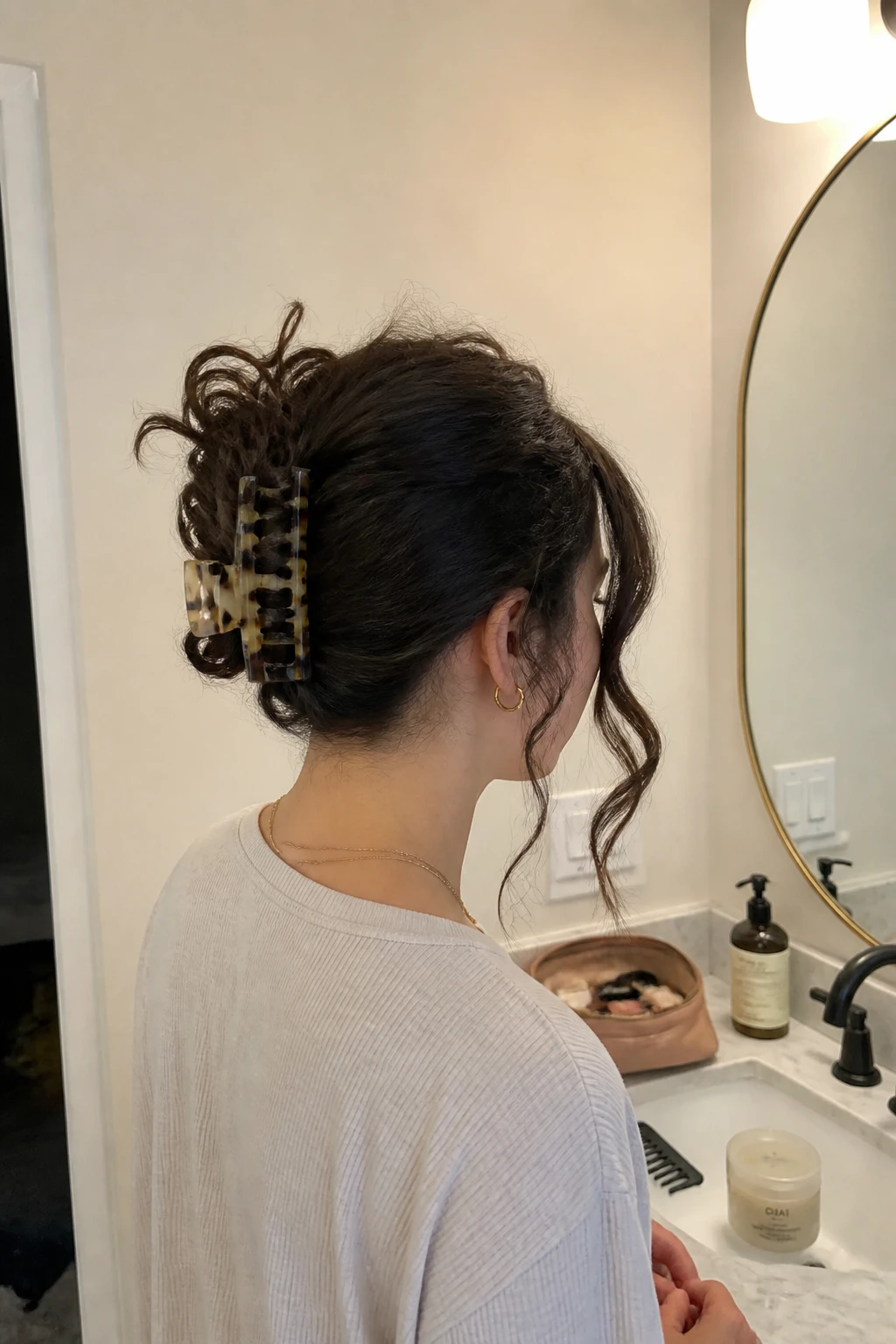 Image prompt

Photo of a woman wearing a loose curly claw clip twist with the hair folded upward, uneven curl ends spilling over a large tortoiseshell clip, and two soft pieces around the face, back three-quarter view beside a bathroom vanity mirror, soft indoor lighting, containing a small lived-in detail such as a wide-tooth comb on the counter, an open makeup bag nearby, and a curl cream jar beside the sink, realistic rushed getting-ready moment, latest iPhone photo quality.