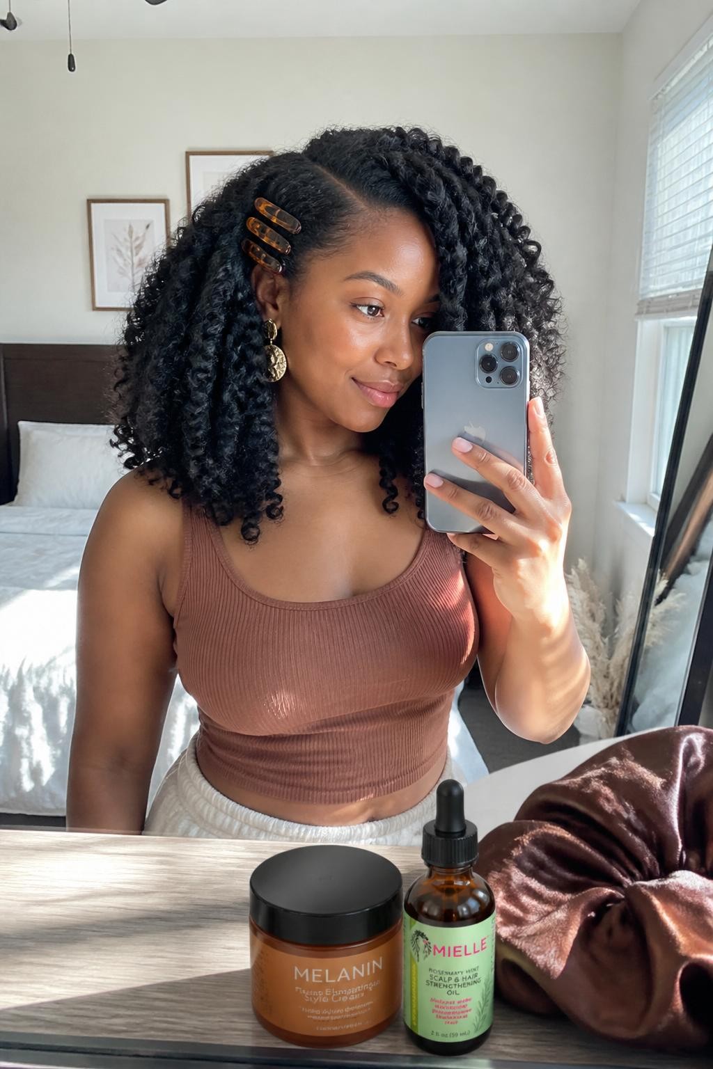 Photo of a Black woman wearing long natural hair in a full defined twist-out with a deep side part, one side pinned above the ear using three amber clips, candid mirror selfie in a sun-drenched bedroom, natural window light, twisting cream and hair oil resting on the dresser, silk bonnet placed nearby and statement earrings peeking through, realistic texture with soft frizz and natural volume, latest iPhone photo quality.