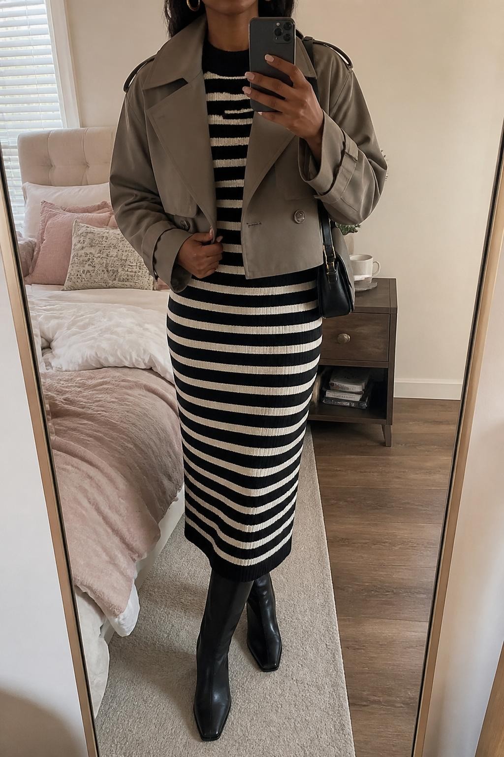 mirror selfie of a Black woman wearing the exact outfit described above, clearly showing a taupe cropped trench jacket layered over a black and cream striped sweater midi dress, black knee-high boots, a black leather shoulder bag, and gold huggie earrings, holding a phone in front of a mirror, cropped at NECK so the face does not show, with a relaxed casual selfie pose, in a feminine-themed bedroom, natural window light, with soft pillows on an unmade bed and a coffee mug on a bedside table, latest iPhone photo quality.