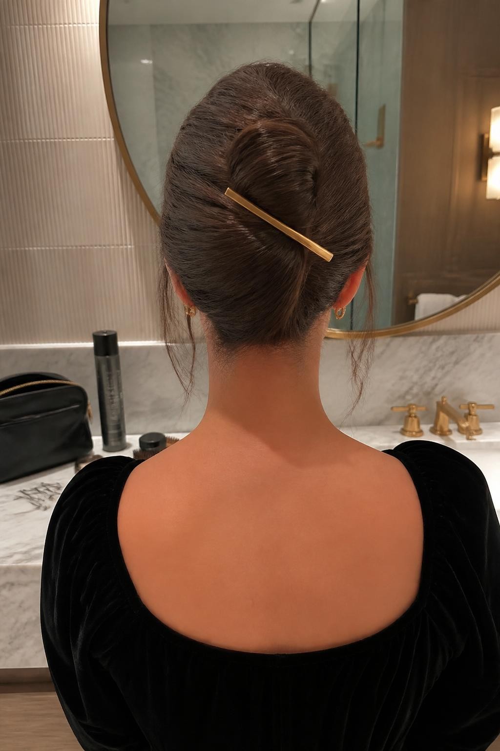 Photo of a woman wearing a softly textured French twist with gentle crown volume, folded ends tucked into the center, and a slim brushed-gold barrette placed diagonally across the twist, back-of-head view in a modern hotel bathroom, soft indoor lighting, texture spray and bobby pins scattered beside a makeup bag, black velvet dress visible at the shoulders and natural loose strands near the ears, latest iPhone photo quality.
