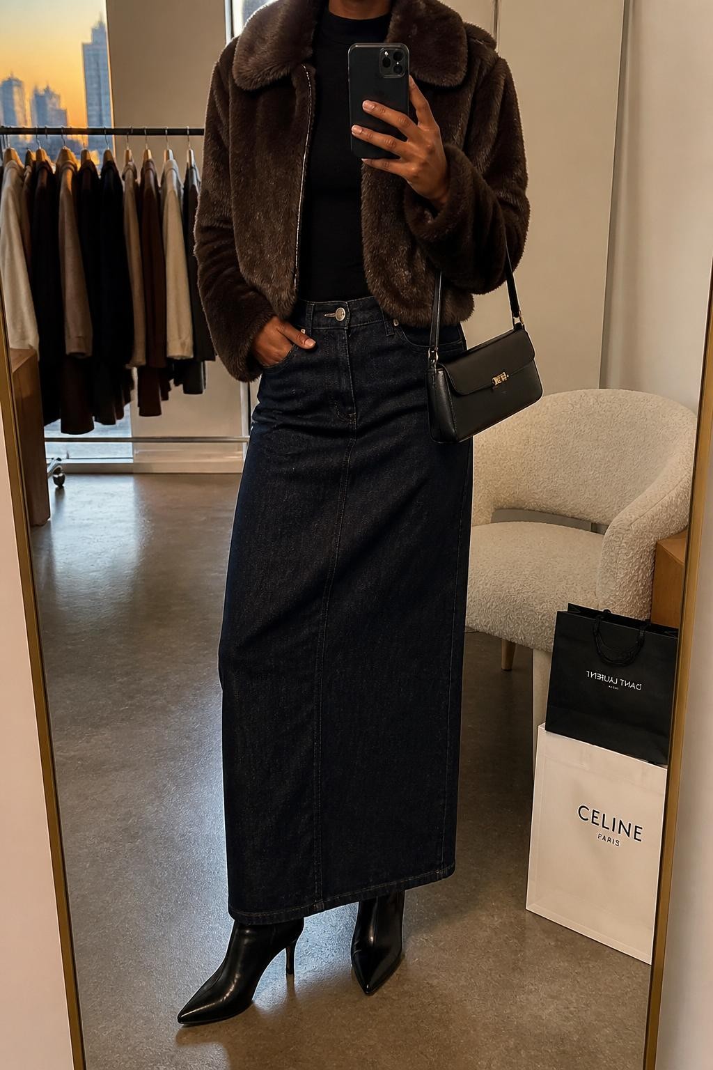 mirror selfie of a Black woman wearing a brown faux fur cropped coat over a black fitted mock-neck top, dark denim maxi skirt, black heeled ankle boots, and a black mini shoulder bag, clearly showing the same clothing items, colors, fabrics, and accessories listed in the Outfit Pieces section, holding a phone in front of a mirror, cropped at NECK so the face does not show, with a relaxed casual selfie pose, in a fashion studio mirror corner, evening golden hour, including a clothing rack with garments behind and shopping bags beside a chair, latest iPhone photo quality.