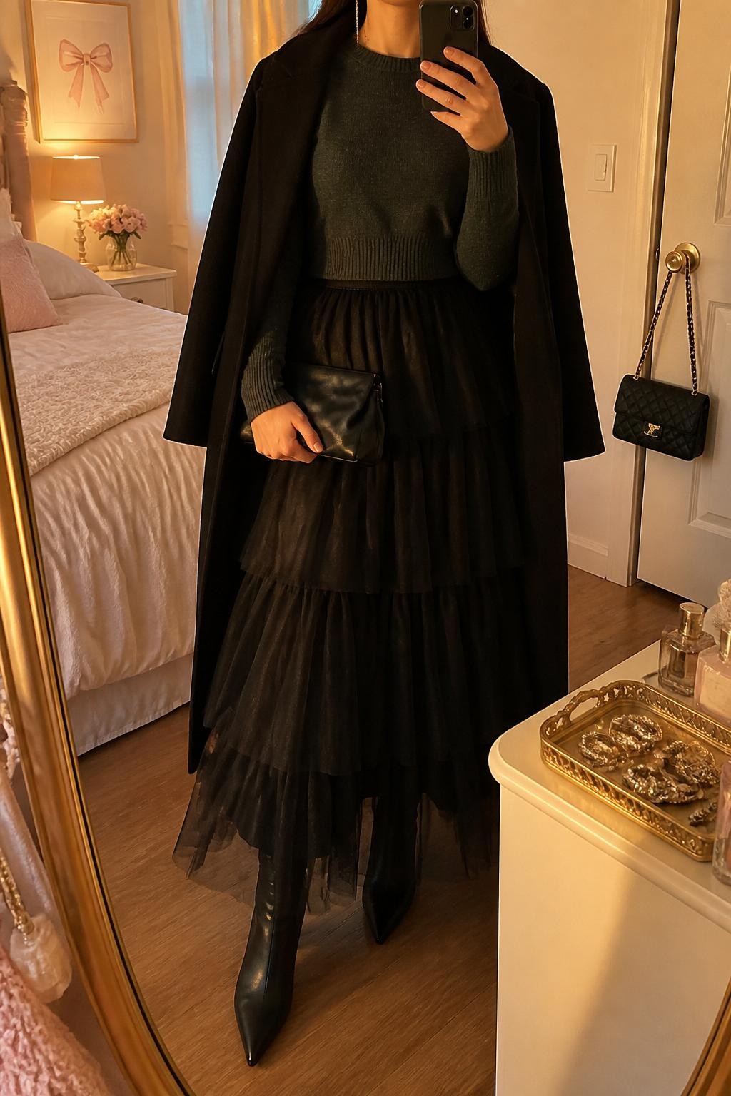mirror selfie of a woman wearing the exact outfit described above, clearly showing a black layered tulle maxi skirt, charcoal cropped knit sweater, black pointed ankle boots, black satin clutch, silver drop earrings, and a black wool coat draped over the shoulders, holding a phone in front of a mirror, cropped at NECK so the face does not show, with a relaxed casual selfie pose, in a feminine-themed bedroom, evening golden hour, with a jewelry tray on the vanity and a handbag hanging on a door handle, latest iPhone photo quality.