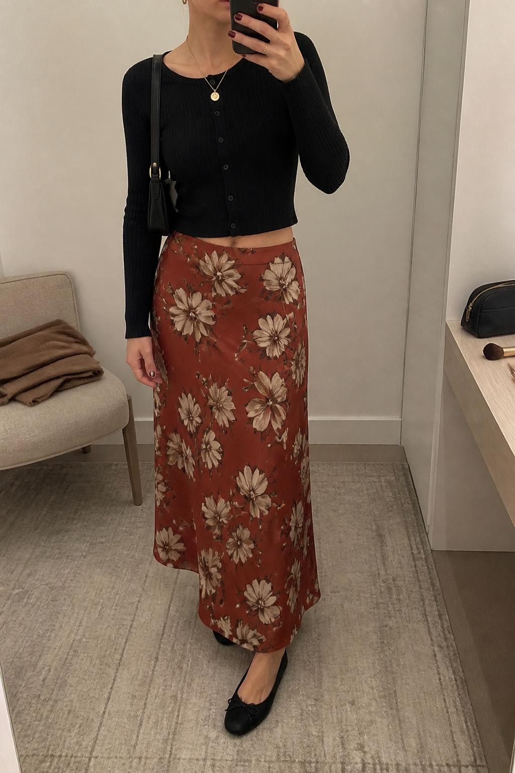 mirror selfie of a woman wearing the exact outfit described above, clearly showing a rust floral maxi skirt with cream and brown flowers, black cropped button-front cardigan, black ballet flats, black mini shoulder bag, gold pendant necklace, and small gold earrings, holding a phone in front of a mirror, cropped at NECK so the face does not show, with a relaxed casual selfie pose, in a minimal dressing room, soft indoor lighting, with a folded blanket on a chair and makeup brush or cosmetic bag on the counter, latest iPhone photo quality.