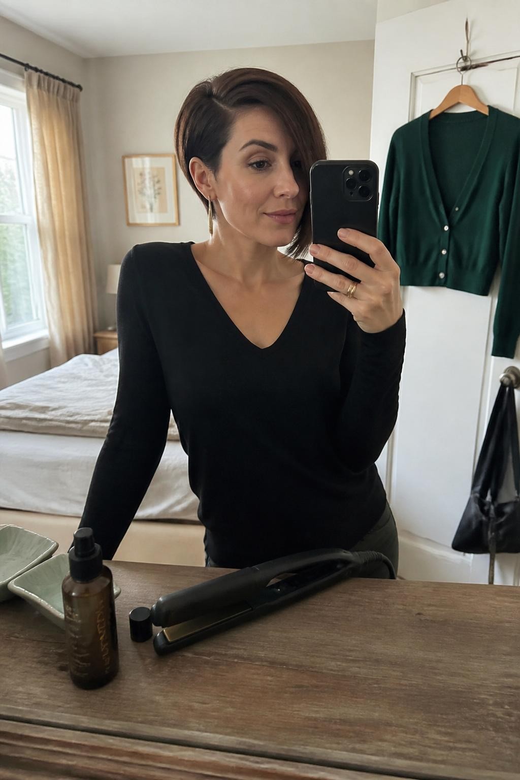 Photo of a woman wearing a short asymmetrical bob with one side grazing the jaw, the opposite side cut above the ear, and a smooth deep side sweep, candid mirror selfie in a softly lit bedroom, warm window light, flat iron and hair serum on the dresser, long drop earring visible on the shorter side and a dark green cardigan hanging nearby, latest iPhone photo quality.