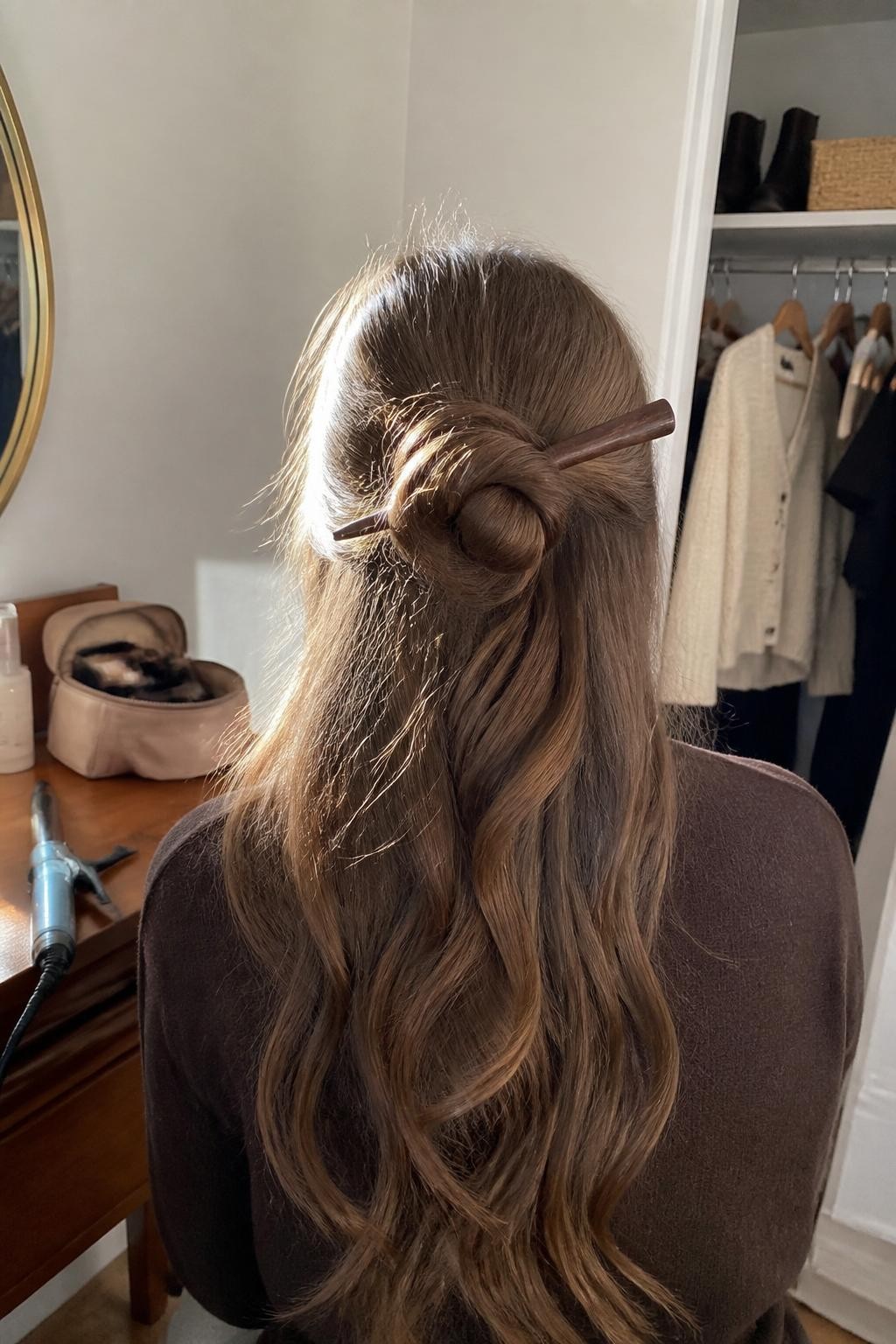 Photo of a woman wearing long softly textured hair with the upper half twisted into a loose knot secured by a slim wooden hair stick, the remaining length falling in natural waves, back-of-head view in a sun-drenched bedroom, warm window light, curling iron on the dresser beside an open makeup pouch, silk scrunchie around her wrist and a cream cardigan visible in a slightly open closet, realistic imperfect knot and flyaways, latest iPhone photo quality.