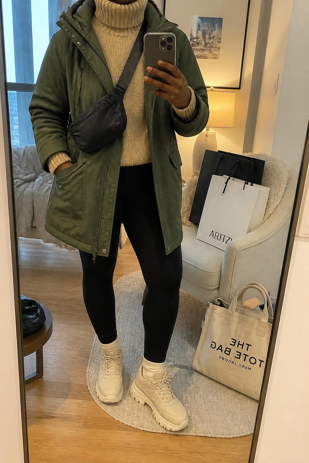 mirror selfie of a Black woman wearing an olive green hooded parka over a beige chunky turtleneck sweater, black fleece-lined leggings, cream winter sneakers, and a black nylon crossbody bag, clearly showing the same clothing items, colors, fabrics, and accessories listed in the Outfit Pieces section, holding a phone in front of a mirror, cropped at NECK so the face does not show, with a relaxed casual selfie pose, in a stylish city apartment, soft indoor lighting, including shopping bags beside a chair and a tote bag resting on the floor, latest iPhone photo quality.