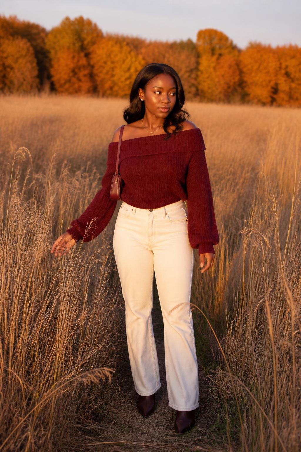 realistic golden-hour senior portrait of a Black young woman wearing a burgundy off-shoulder knit sweater, ecru high-waisted straight-leg jeans, chocolate brown pointed ankle boots, a thin gold necklace, and a burgundy mini shoulder bag, standing in a field of tall dry grass with distant orange trees, full-body portrait clearly showing the exact clothing colors, knit texture, denim, boots, and accessories, natural pose with one hand brushing the grass, warm sunset lighting, realistic skin and fabric texture, professional DSLR quality.