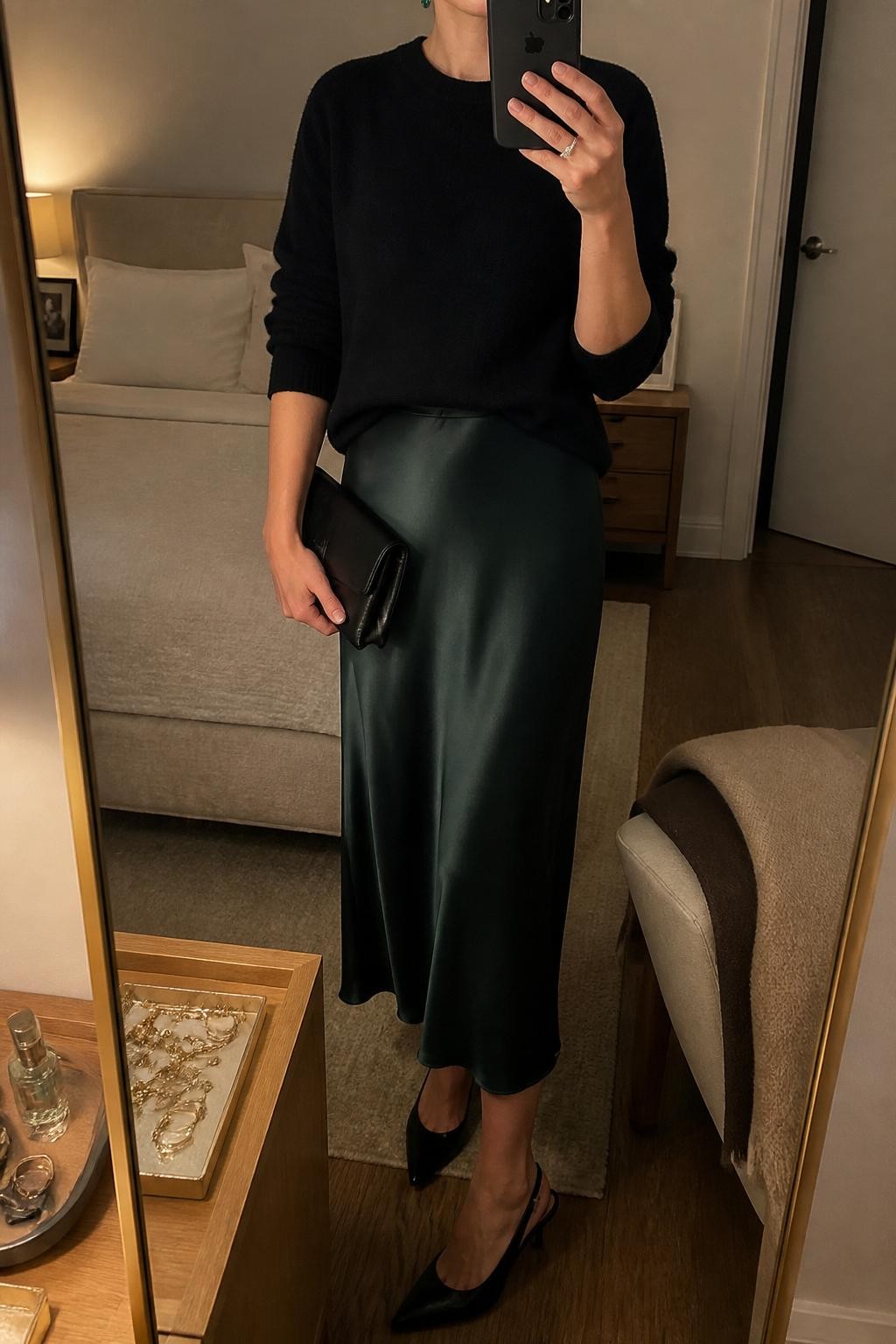 mirror selfie of a woman wearing the exact outfit described above, clearly showing a black cashmere sweater, forest green satin slip skirt, black slingback heels, emerald stud earrings, and a black clutch, holding a phone in front of a mirror, cropped at NECK so the face does not show, with a relaxed casual selfie pose, in a cozy bedroom mirror setup, warm moody lighting, with a folded blanket on a chair and a jewelry tray on the vanity, latest iPhone photo quality.