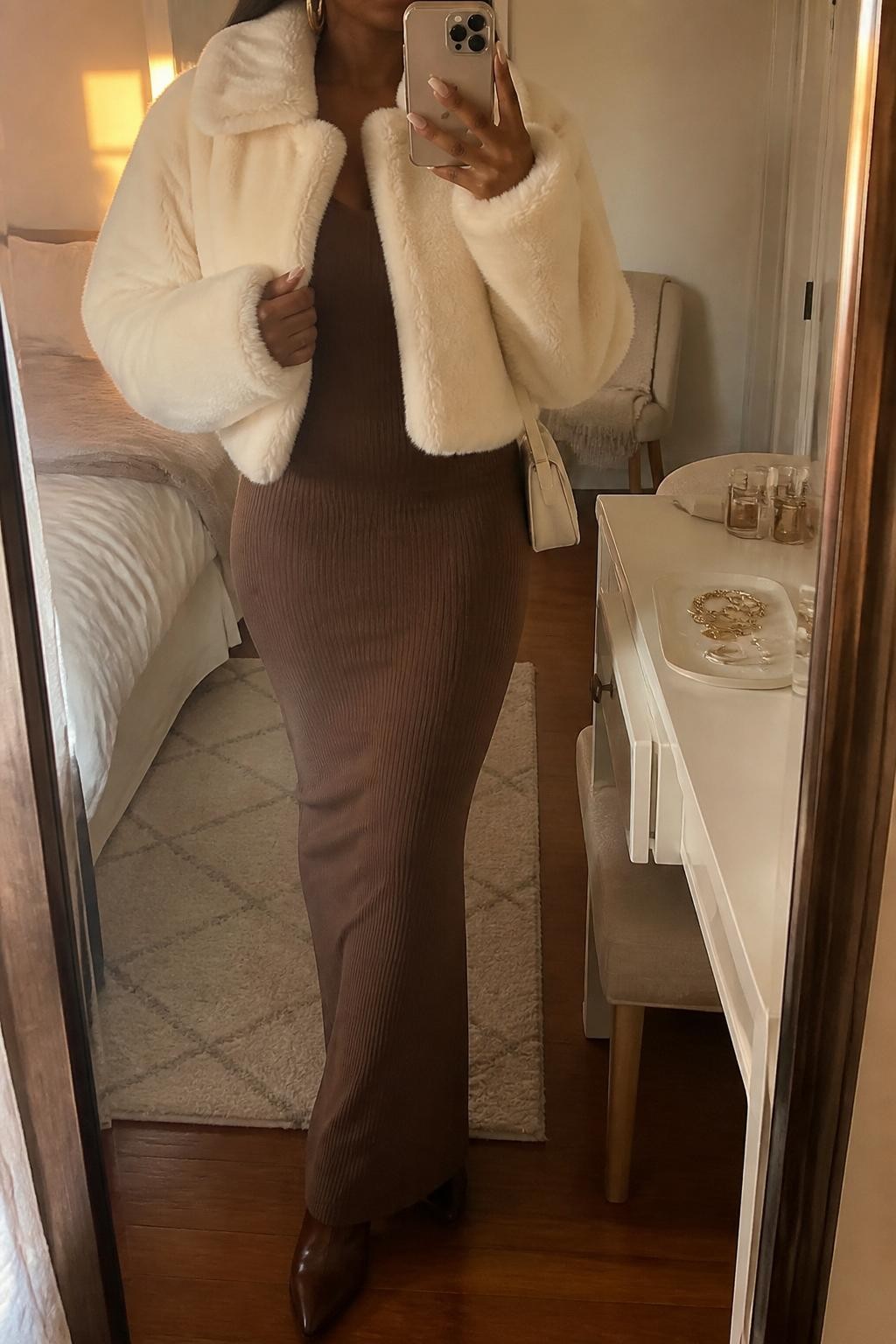 mirror selfie of a Black woman wearing the exact outfit described above, clearly showing a brown ribbed maxi dress, cropped cream faux fur jacket, brown heeled boots, gold hoops, and a cream mini shoulder bag, holding a phone in front of a mirror, cropped at NECK so the face does not show, with a relaxed casual selfie pose, in a cozy bedroom mirror setup, evening golden hour, with a folded blanket on a chair and a jewelry tray on the vanity, latest iPhone photo quality.