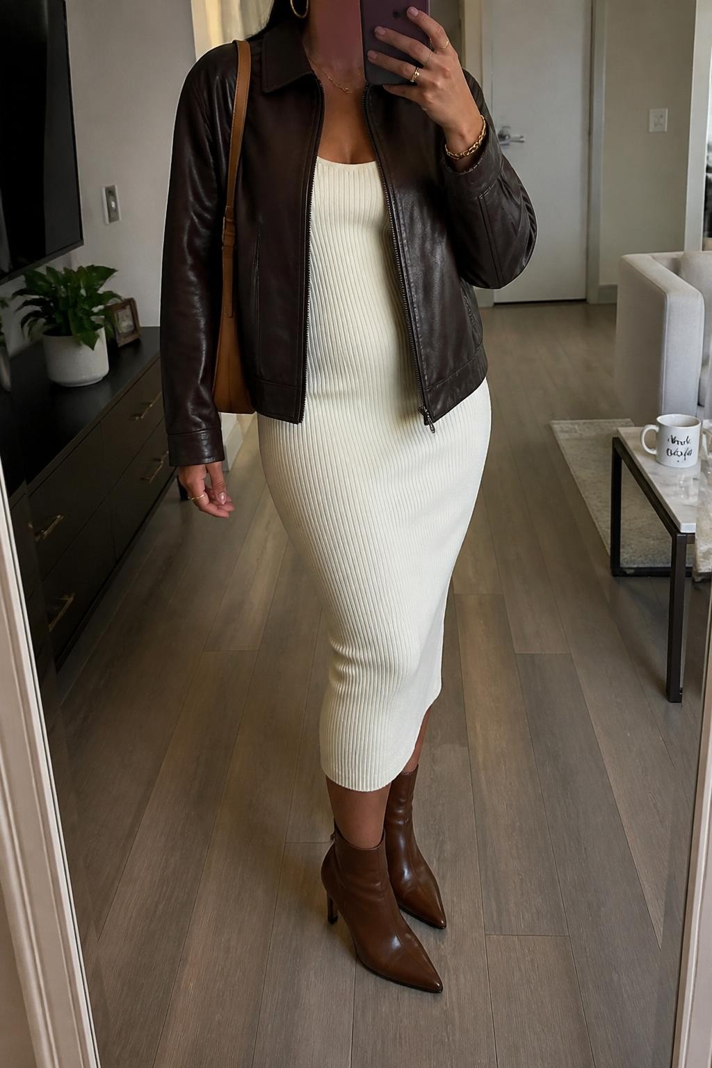 mirror selfie of a Latina woman wearing a chocolate brown leather jacket over a cream ribbed knit midi dress, brown heeled ankle boots, gold hoop earrings, and a tan shoulder bag, holding a phone in front of a mirror, cropped at NECK so the face does not show, relaxed casual selfie pose, in a stylish city apartment with soft indoor lighting, including a coffee mug on a side table and a small plant on a dresser, realistic latest iPhone photo quality.