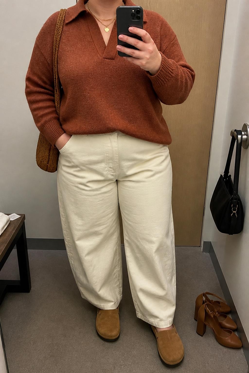 mirror selfie of a plus size woman wearing the exact outfit described above, a warm rust knit polo sweater with a soft collar, cream cotton twill barrel pants, tan suede clogs with rounded toes, light brown woven shoulder bag, and gold layered necklace with a simple charm, clearly showing the same clothing items, colors, fabrics, and accessories listed in the Outfit Pieces section, holding a phone in front of a mirror, cropped at NECK so the face does not show, with a relaxed casual selfie pose, in a minimal dressing room, soft indoor lighting, including a handbag hanging on a door handle and a pair of heels on the floor nearby, latest iPhone photo quality.
