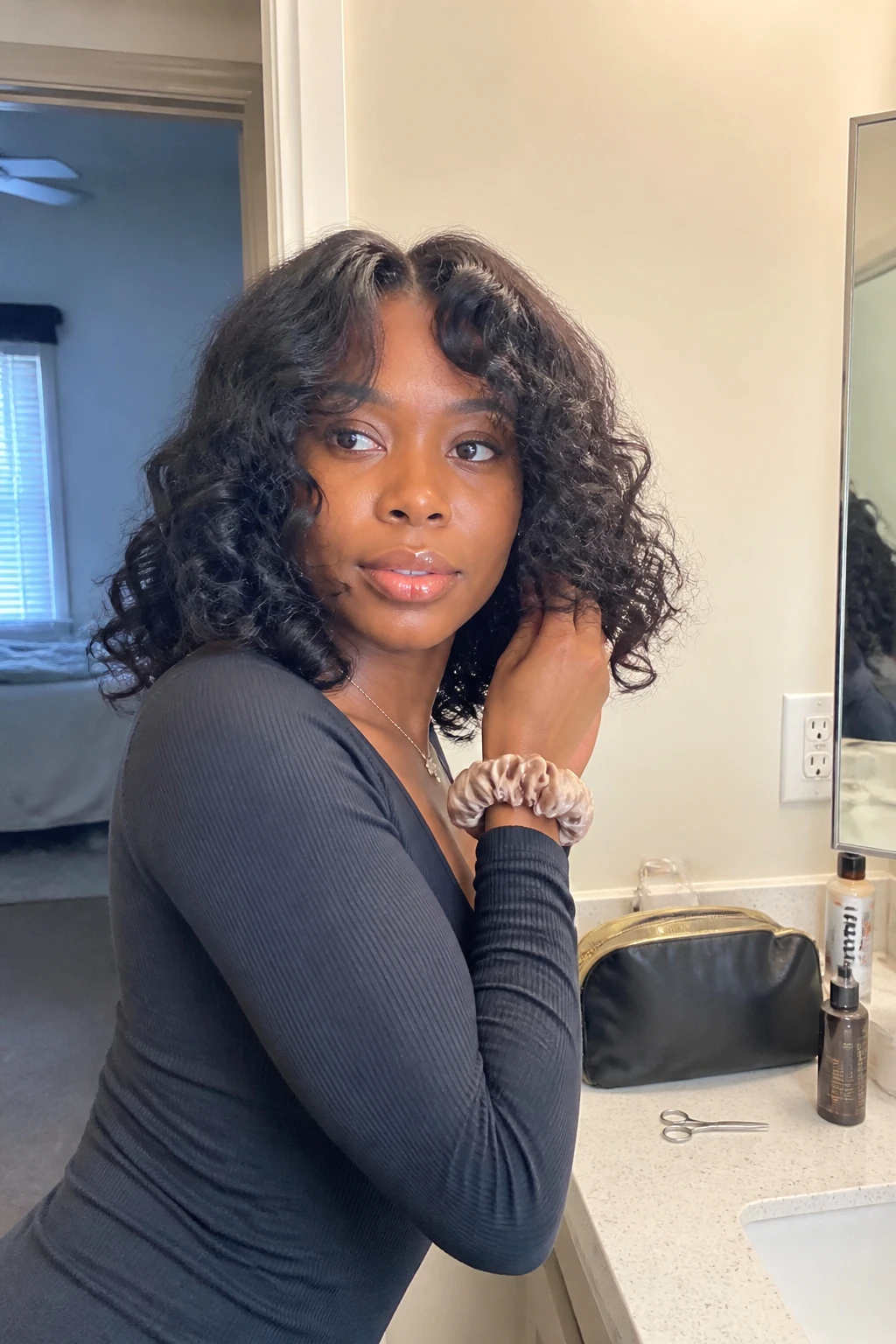 Image prompt
Photo of a Black woman wearing a medium-length curly sew-in with soft curtain bangs opening around the forehead, defined curls framing the cheeks, and light volume through the sides, front three-quarter view beside a bathroom vanity mirror, natural window light, containing a small lived-in detail such as small trimming scissors and curl foam on the counter, a makeup bag nearby, and a silk scrunchie on her wrist, realistic getting-ready moment, latest iPhone photo quality.
