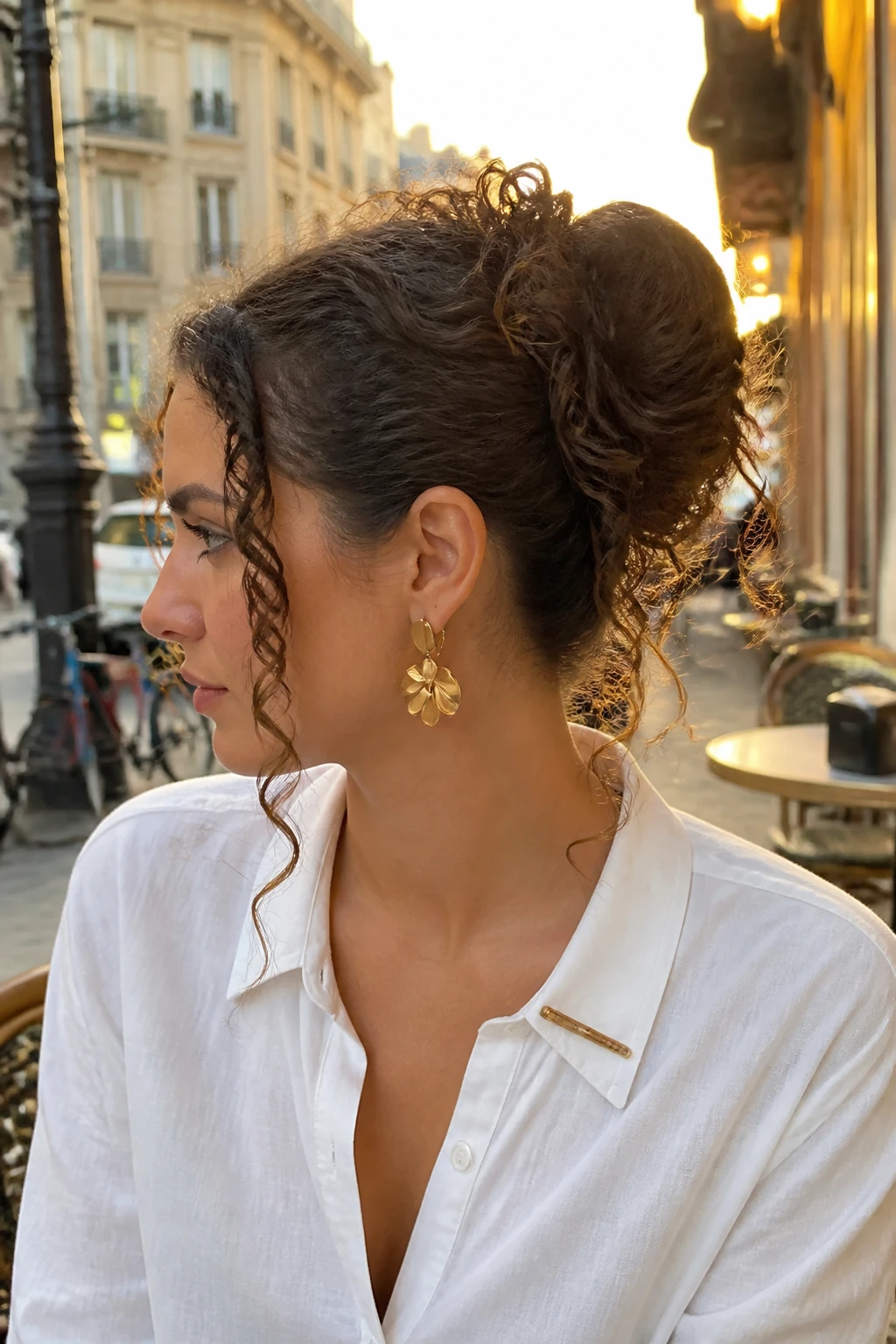 Image prompt

Photo of a woman wearing a curly ponytail bun hybrid with a rounded pinned base and several defined curls falling freely from the centre, side profile view highlighting the mixed shape, softly blurred city street background, golden hour glow, containing a small lived-in detail such as statement earrings and a slim hair clip clipped to her collar, realistic candid photograph, latest iPhone photo quality.