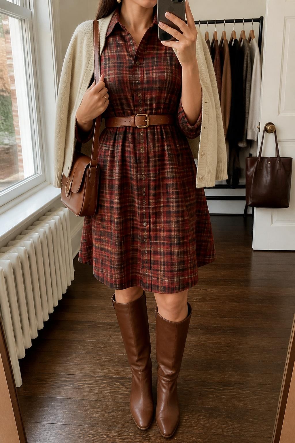 mirror selfie of a woman wearing the exact outfit described above, clearly showing chestnut brown tall boots, a red and brown plaid button-front shirt dress, a tan waist belt, a cream cardigan draped over the shoulders, a brown satchel bag, and gold stud earrings, holding a phone in front of a mirror, cropped at NECK so the face does not show, with a relaxed casual selfie pose, in a fashion studio mirror corner, natural window light, with clothing rack with garments behind and a handbag hanging on a door handle, latest iPhone photo quality.