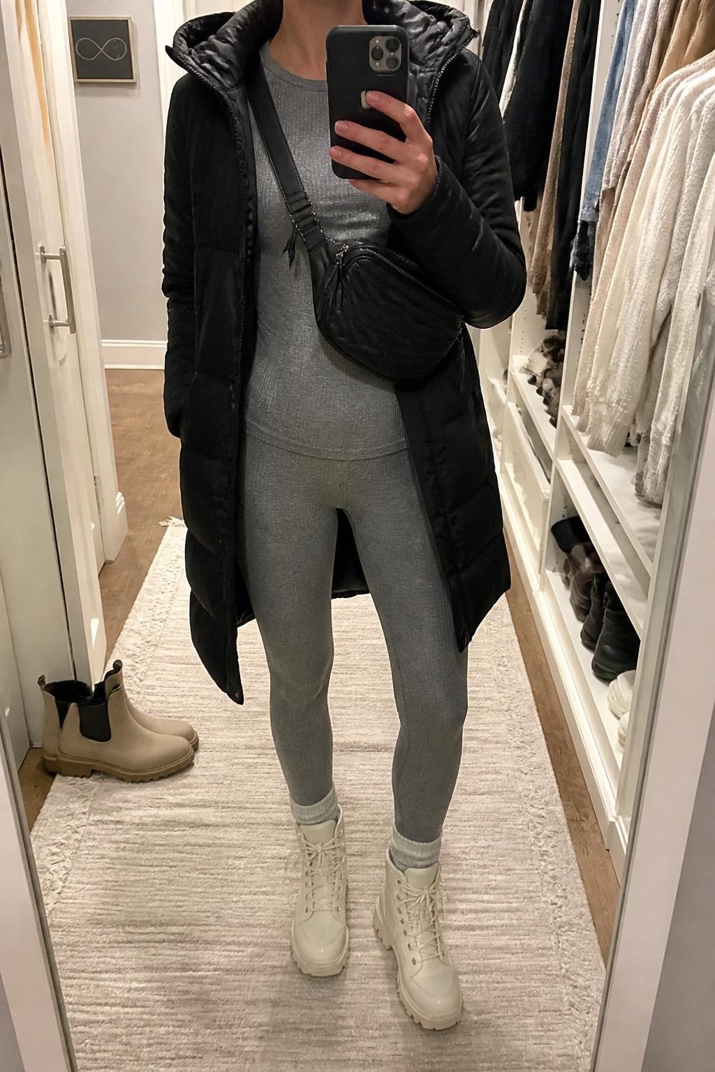 mirror selfie of a woman wearing the exact outfit described above, clearly showing a black longline puffer coat, matching gray thermal long-sleeve top and gray thermal leggings, cream hiking boots, and a black quilted belt bag worn crossbody, holding a phone in front of a mirror, cropped at NECK so the face does not show, with a relaxed casual selfie pose, in a walk-in closet mirror, soft indoor lighting, with an open closet with clothes visible and a pair of boots on the floor nearby, latest iPhone photo quality.