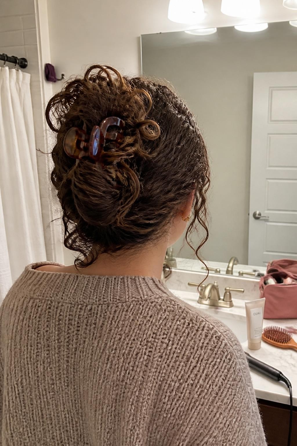 Photo of a woman wearing long curly hair folded into a loose French twist and held with an oversized tortoiseshell claw clip, several curled ends spilling over the top and soft tendrils near the ears, back-of-head view at a bathroom vanity mirror, warm indoor lighting, curl cream and hairbrush on the counter beside an open makeup bag, knit sweater visible and a curling iron left nearby, realistic imperfect getting-ready moment, latest iPhone photo quality.