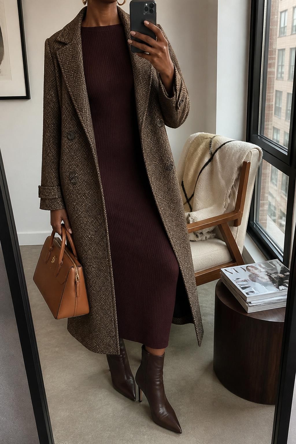 mirror selfie of a Black woman wearing a long brown herringbone wool coat, dark burgundy fitted knit midi dress, chocolate brown heeled ankle boots, gold hoop earrings, and a structured cognac leather handbag, clearly showing the same clothing items, colors, fabrics, and accessories listed in the Outfit Pieces section, holding a phone in front of a mirror, cropped at NECK so the face does not show, with a relaxed casual selfie pose, in a stylish city apartment, natural window light, including a folded blanket on a chair and a stack of fashion magazines on a side table, latest iPhone photo quality.