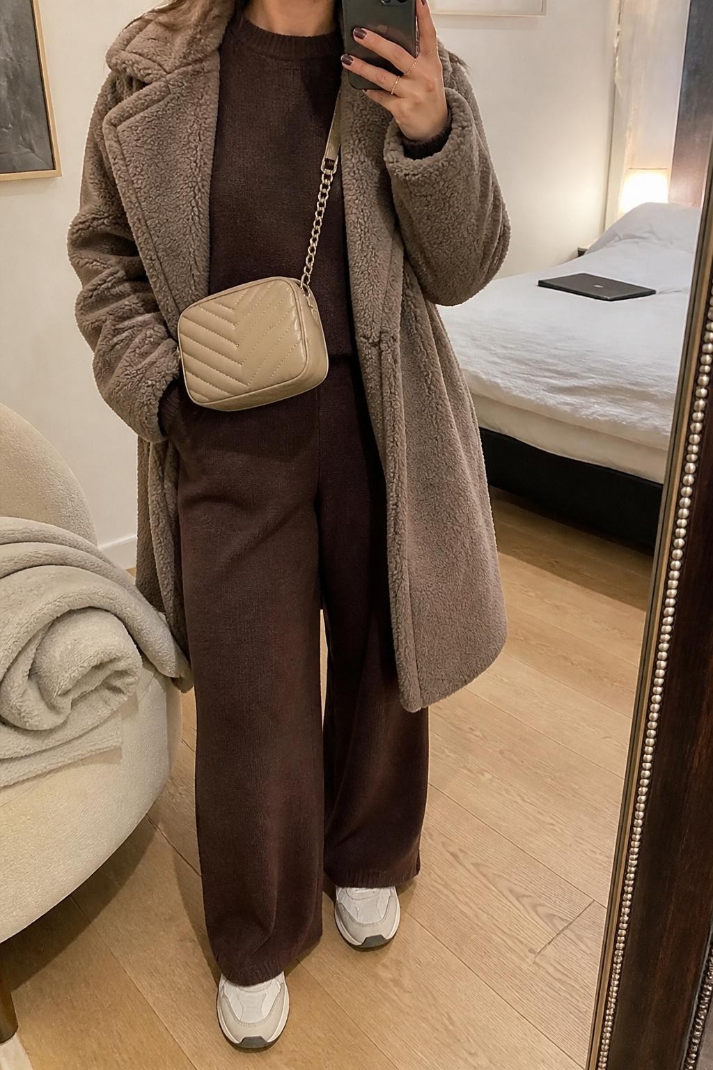 mirror selfie of a woman wearing the exact outfit described above, clearly showing a taupe teddy coat, chocolate brown knit sweater, matching chocolate knit wide-leg pants, cream sneakers, and a beige quilted crossbody bag, holding a phone in front of a mirror, cropped at NECK so the face does not show, with a relaxed casual selfie pose, in a cozy bedroom mirror setup, soft indoor lighting, with a folded blanket on a chair and a laptop or tablet on the bed, latest iPhone photo quality.